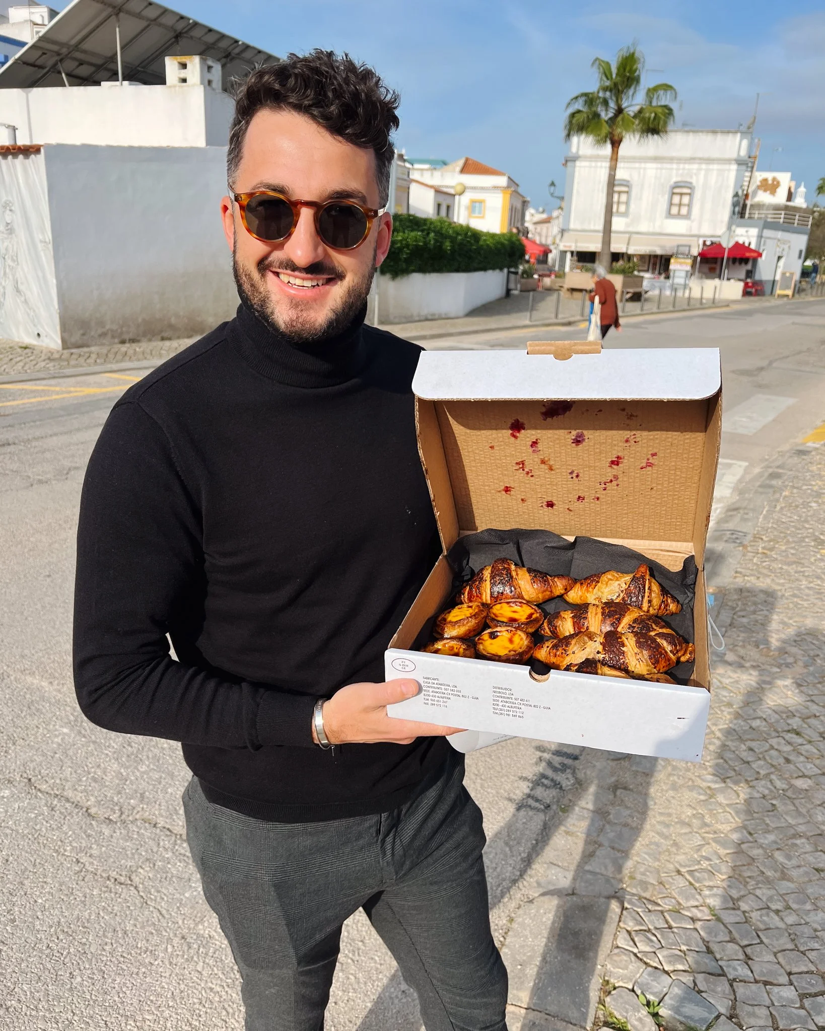 A man in sunglasses, black turtleneck, and gray pants holding an open box of assorted pastries, standing on a city street.
