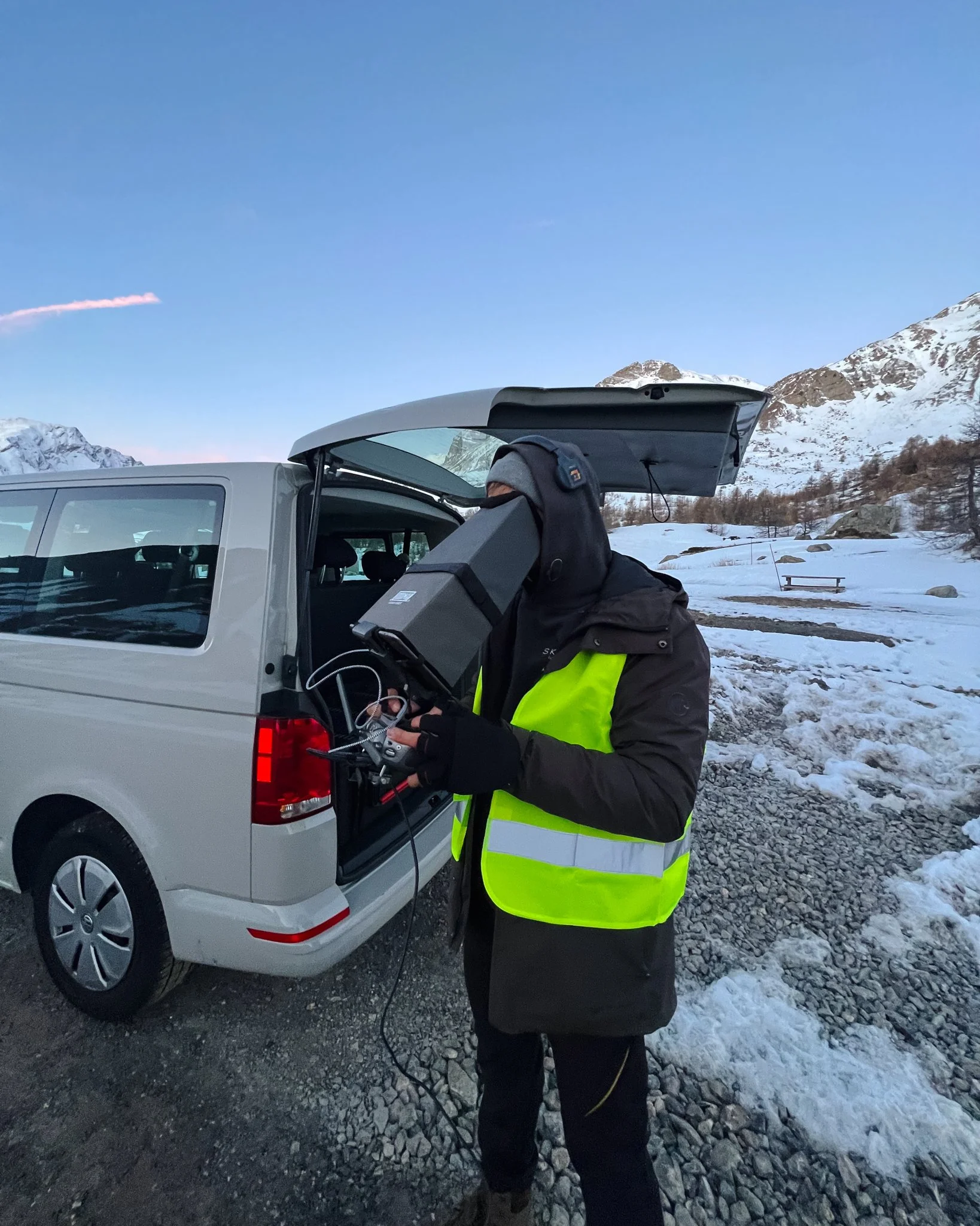 Person in a yellow safety vest and winter clothing operating scientific equipment from the back of a silver van in a snowy mountain landscape.
