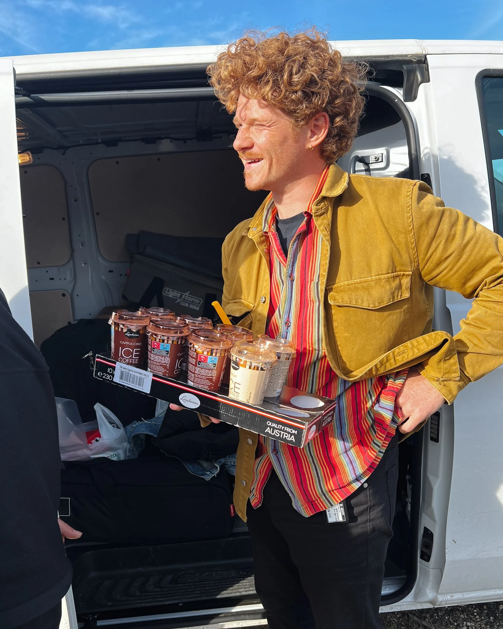 A man with curly hair loading cups of coffee from a tray into a white van, wearing a striped shirt and yellow jacket, outdoors under a blue sky.