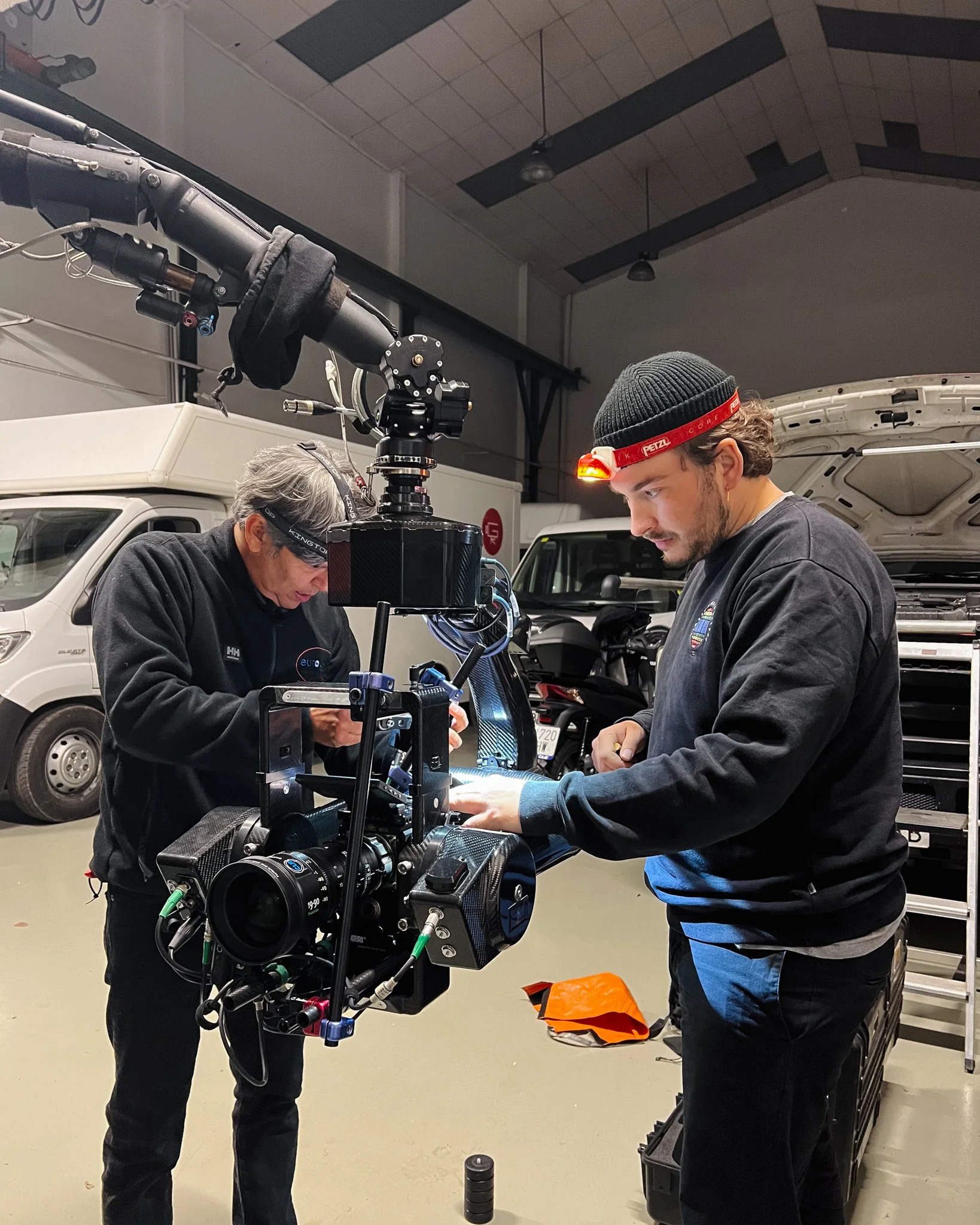 Two men working with a professional camera mounted on a rig in a garage-like space, surrounded by vehicles and equipment