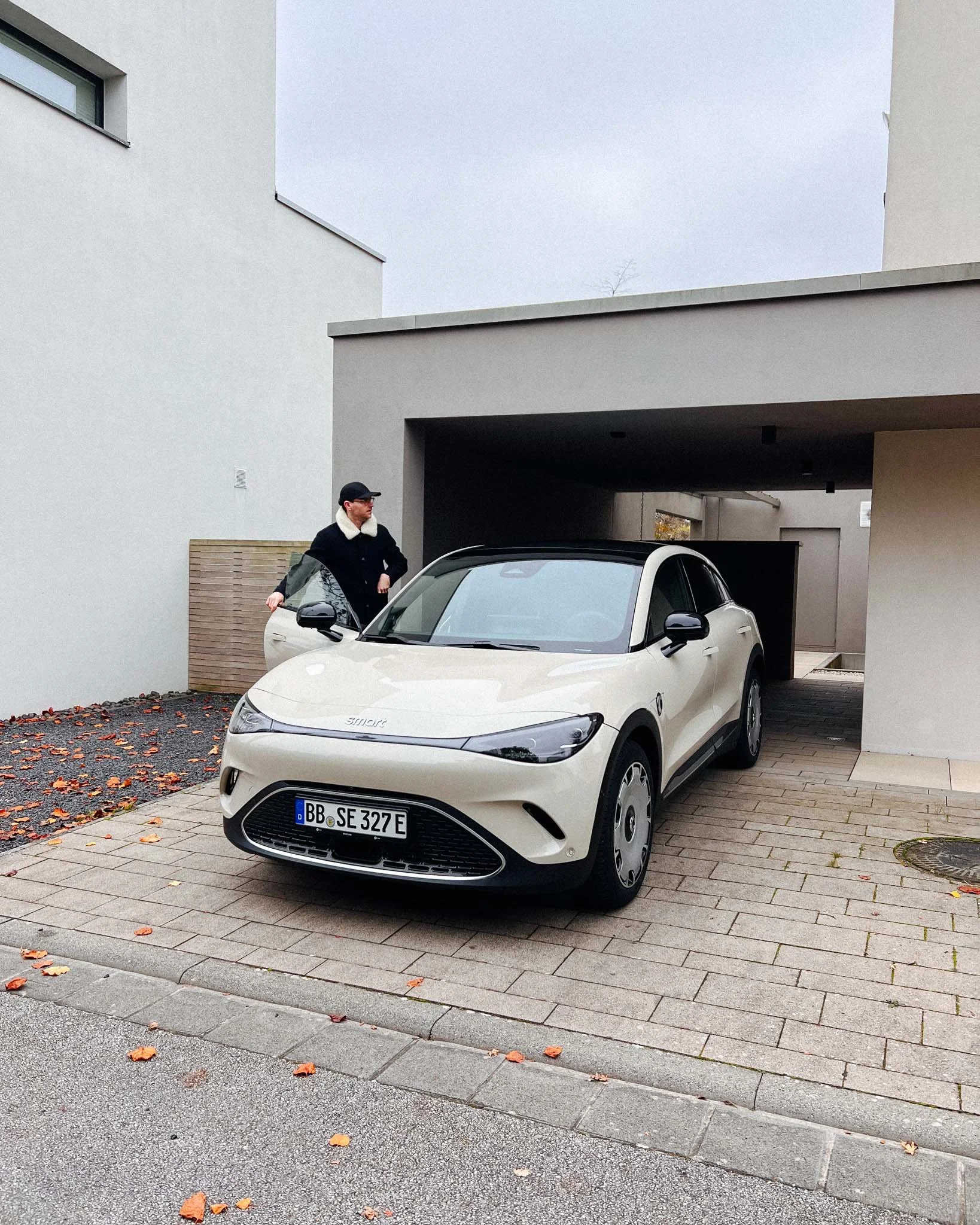 A person in a black coat and cap standing next to a white electric car parked in a modern residential area with gray buildings and fallen autumn leaves on the ground.