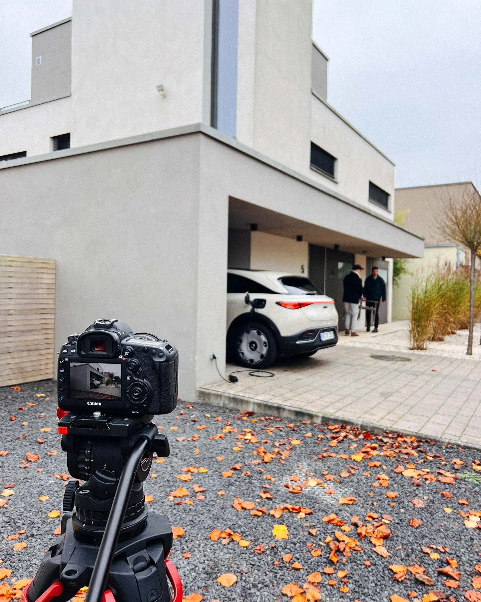 A camera on a tripod captures a modern house with two people standing outside, near a white electric car charging station, on a gravel area scattered with orange and yellow fallen leaves.
