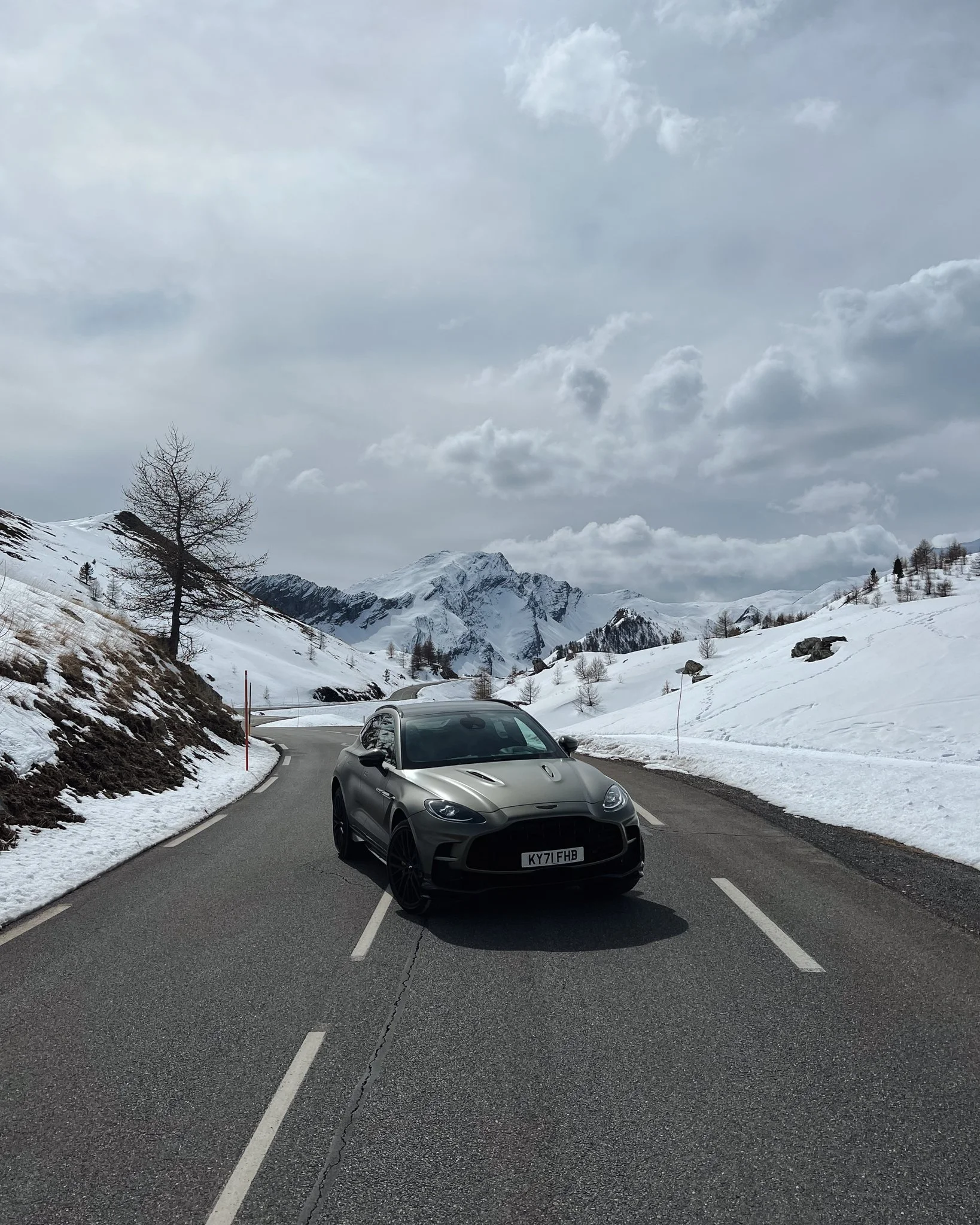 A black sports car parked on a winding mountain road surrounded by snow-covered slopes and mountains under a cloudy sky.