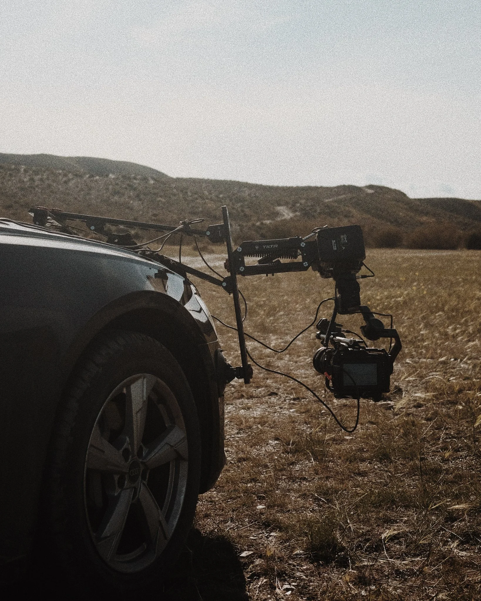 Camera mounted on a car's front bumper in an open field with rolling hills in the background.