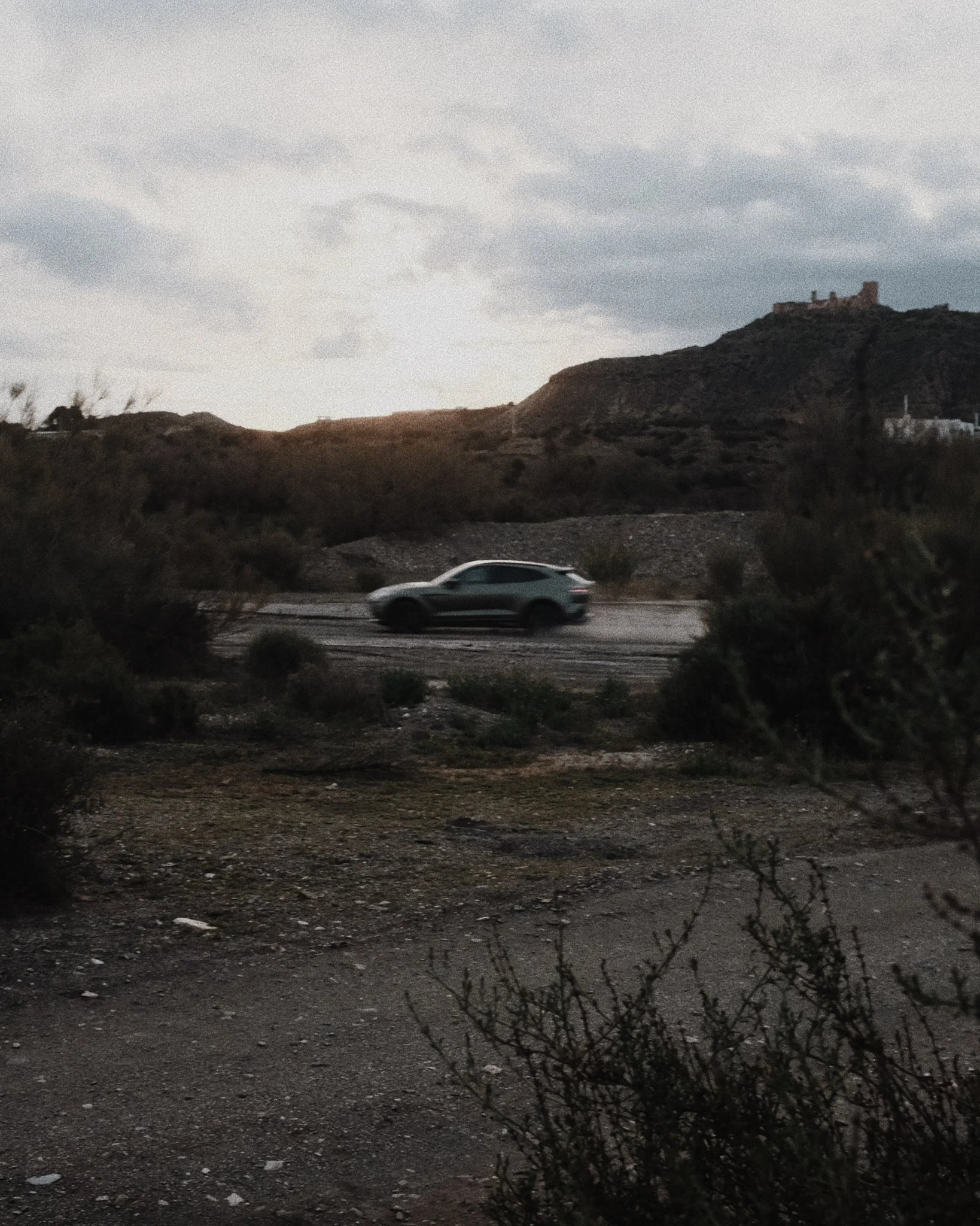 A moving gray SUV on a dirt road in a desert landscape with shrubs, hills, and a castle on a hill in the background under a cloudy sky.