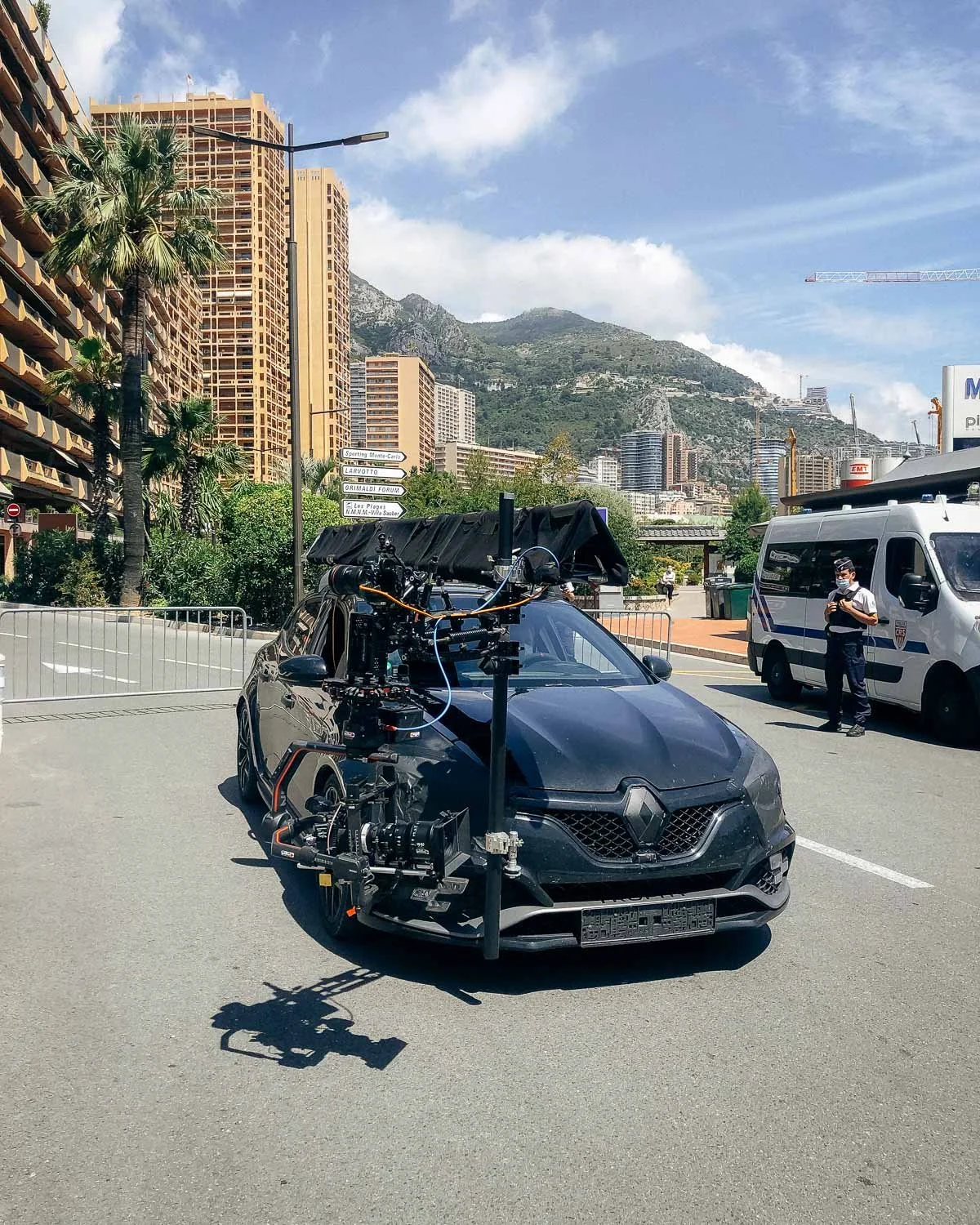 A black Renault car equipped with a mounted camera rig for filming or capturing footage, parked in an urban city area with buildings, trees, and mountains in the background, along with a police officer and vehicle nearby.