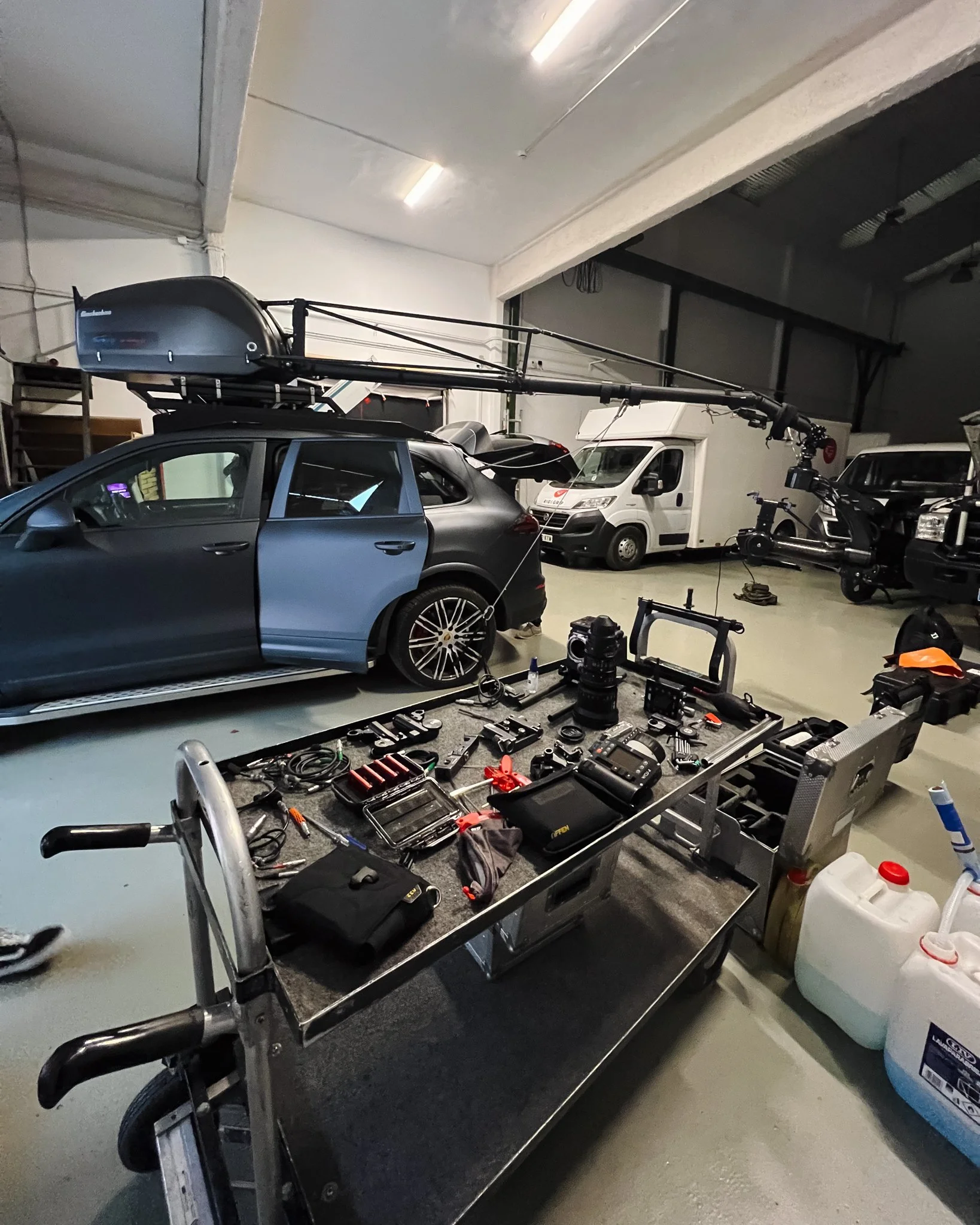 A workshop with a matte gray station wagon that has a large black rooftop cargo box and is equipped with a robotic crane arm attached to it. The workspace includes a workbench with various tools and equipment, and additional vehicles and equipment in