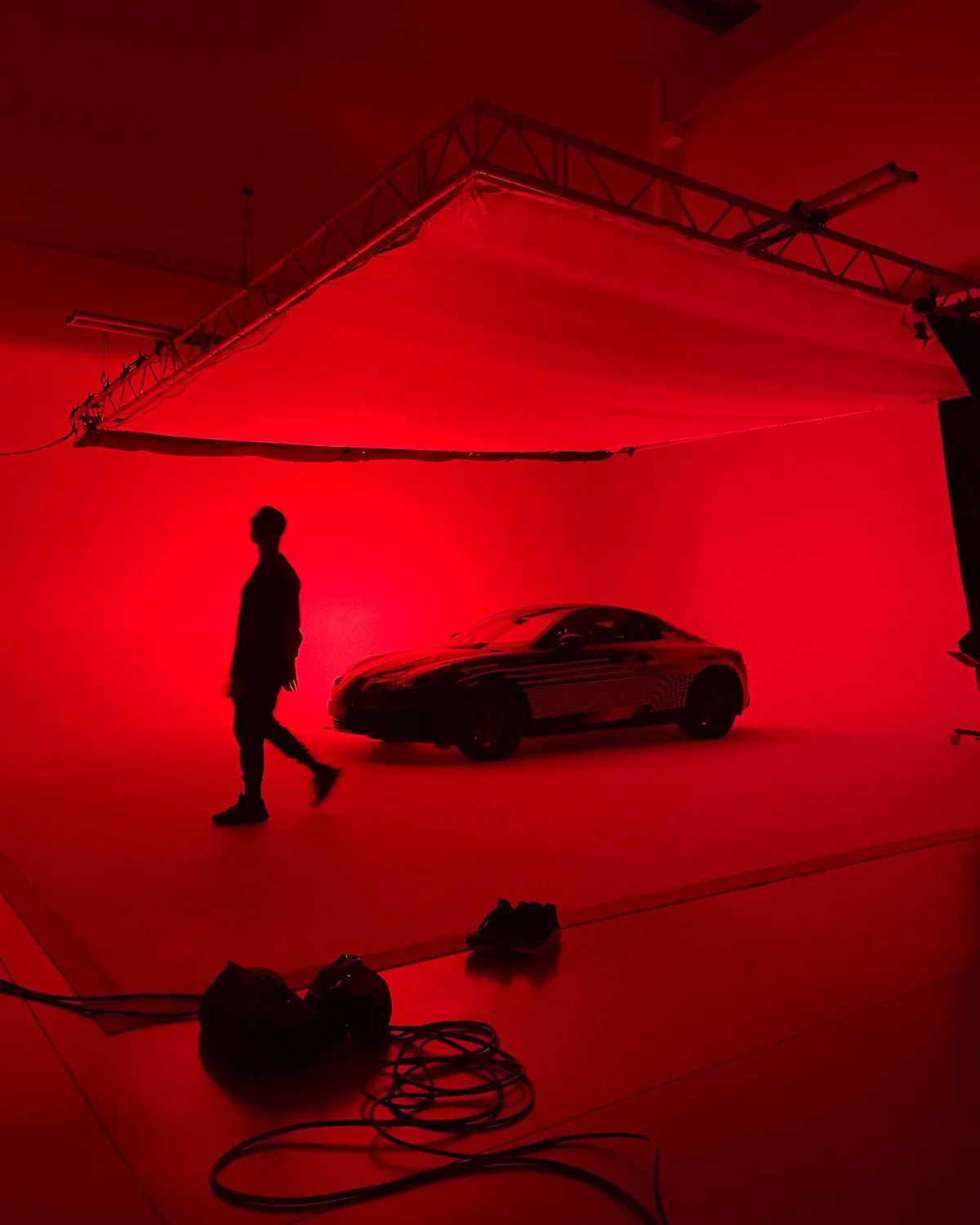 A person walking in a photo studio lit with red lighting, with a sleek black sports car and studio equipment surrounding them.
