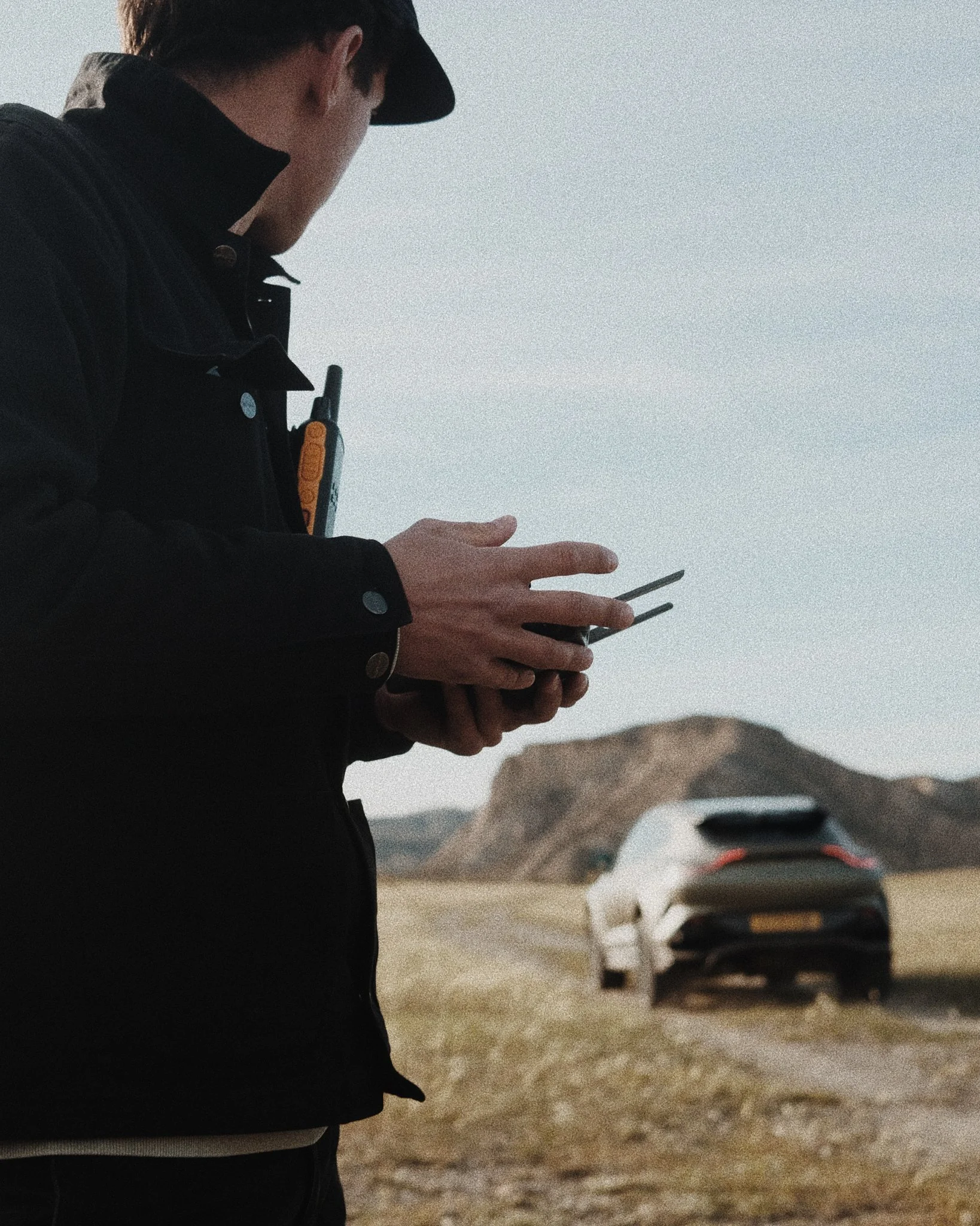 A person in a black jacket and cap using a remote control, with a landscape and mountains in the background and a white car parked on a dirt road.
