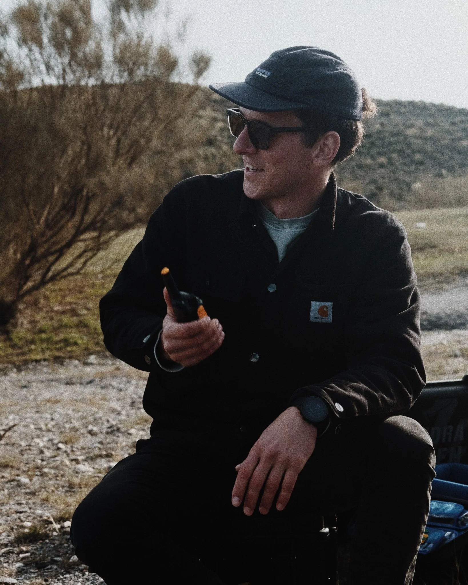A young man wearing sunglasses, a cap, and a Carhartt jacket, sitting outdoors on rocky ground holding a walkie-talkie.