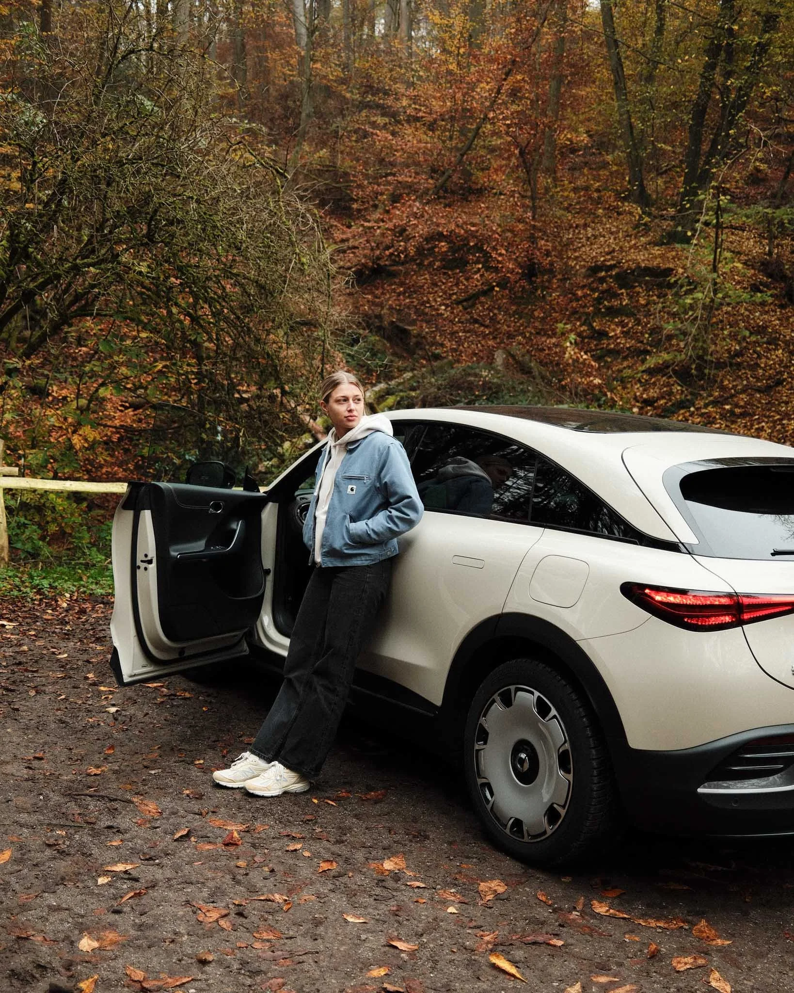 A young woman leaning against a white SUV parked on a forest dirt road during autumn, surrounded by fall foliage with orange and brown leaves.