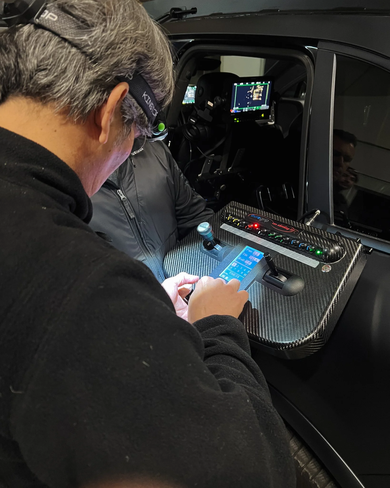 Person using a smartphone near a racing simulator setup with a carbon fiber surface, gear shift, and control panel inside a car.