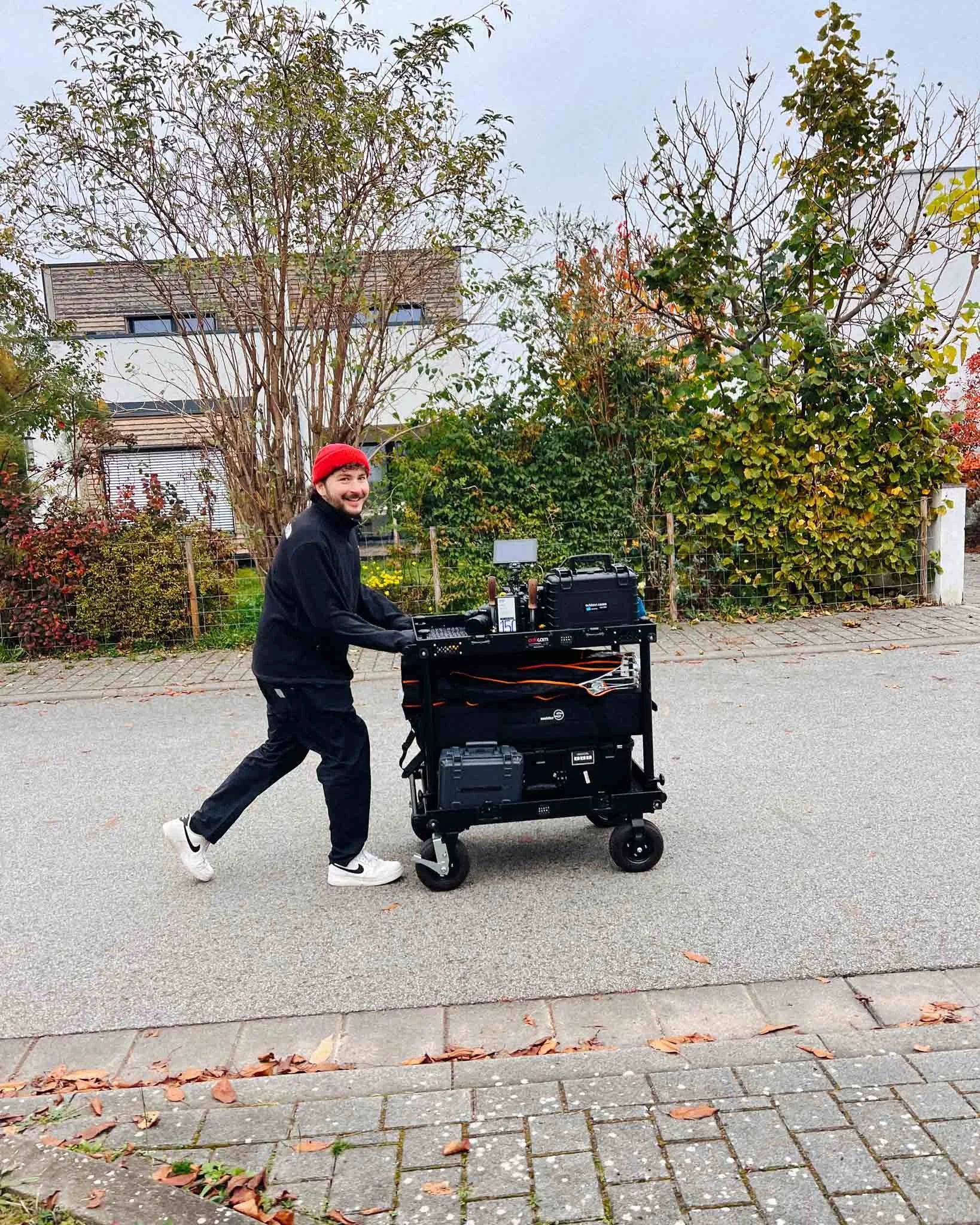 A man in a red beanie, black jacket, and white sneakers pushing a cart with electronic equipment on a residential street with trees and bushes in the background.