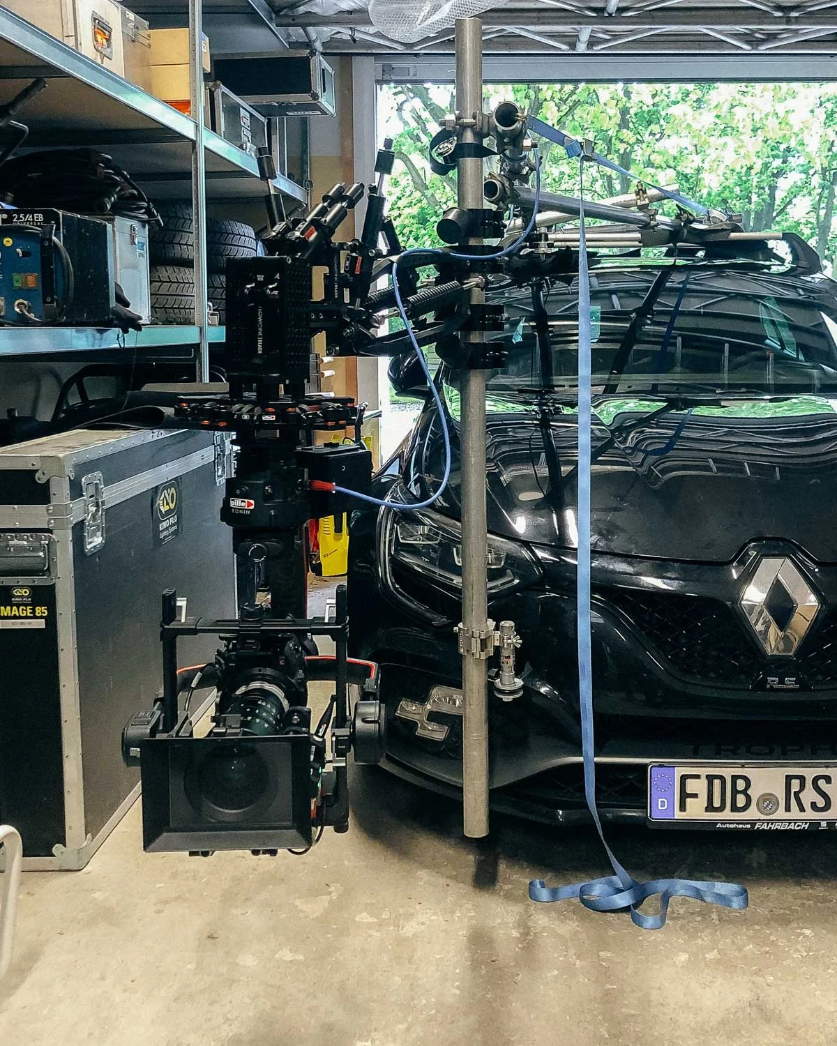 A professional camera mounted on a rig facing a black Renault car in a garage, with equipment and tires stored on shelves in the background.