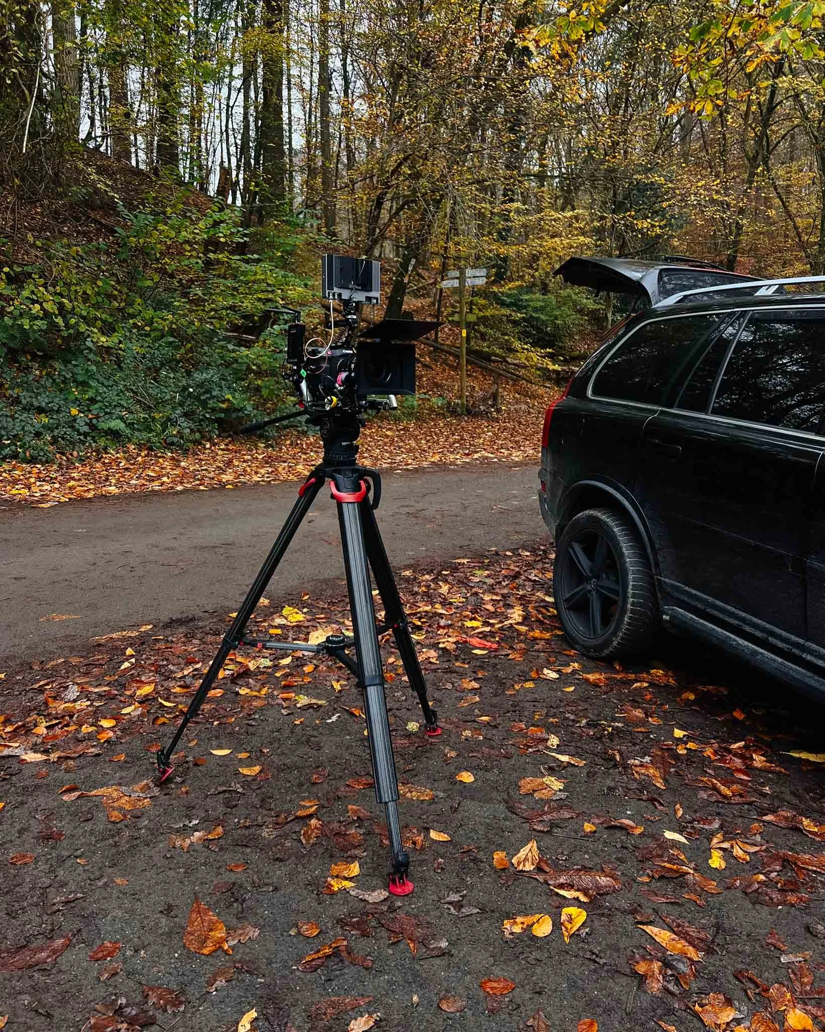 A camera on a tripod set up outdoors on a dirt road covered with fallen autumn leaves, with a black vehicle nearby, in a wooded area with trees showing fall foliage.