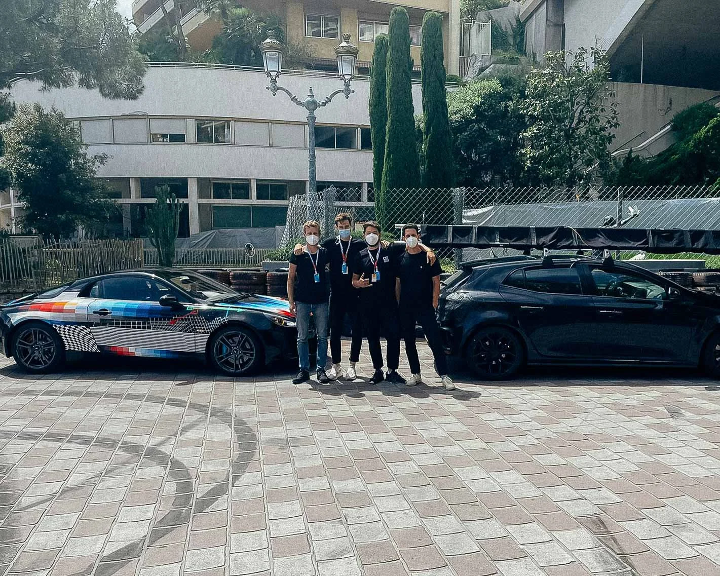 Four people wearing masks and lanyards standing between two race cars in an outdoor area with buildings and trees in the background.
