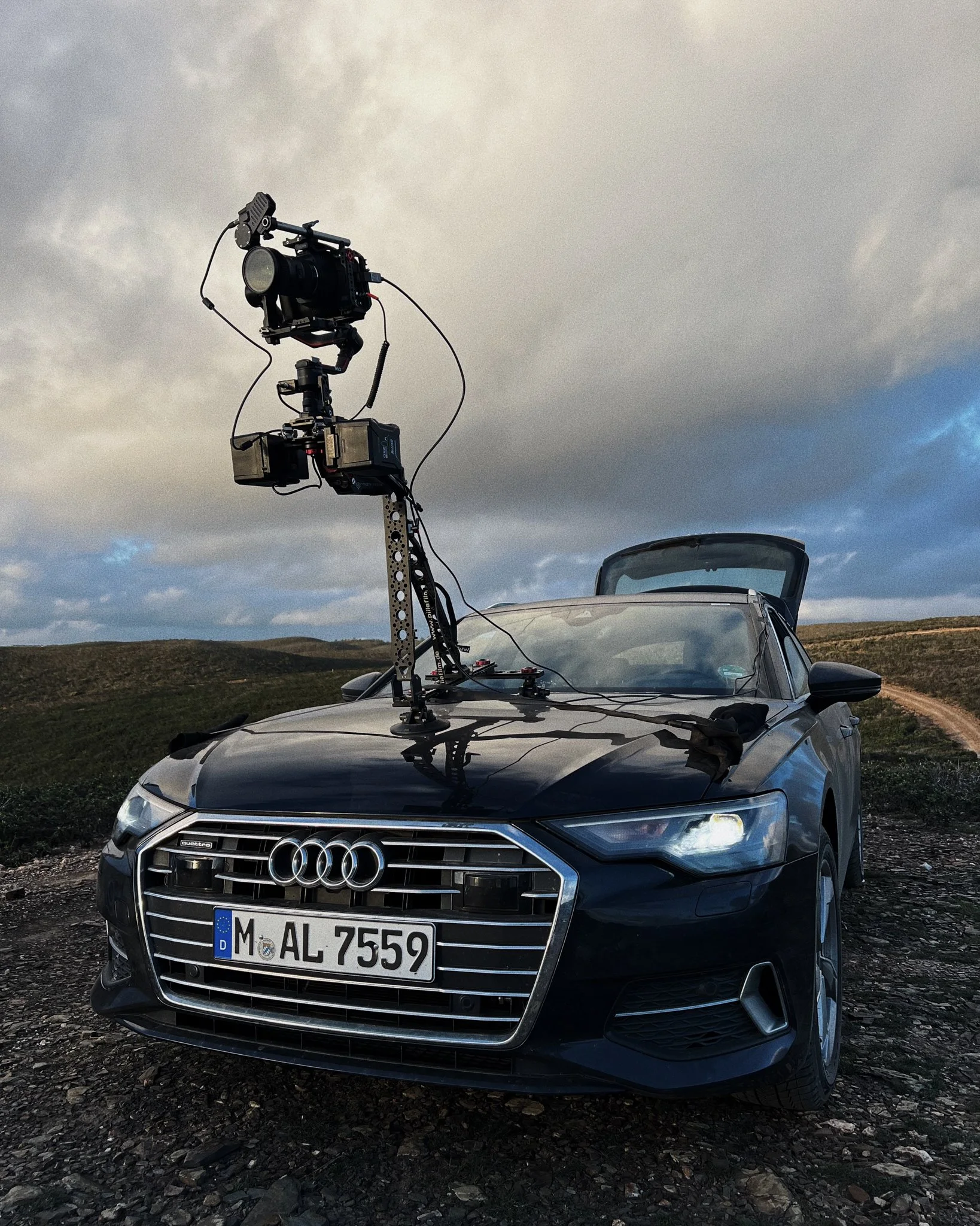 Black Audi car with a mounted camera rig on its hood, set outdoors on a dirt road with cloudy skies.