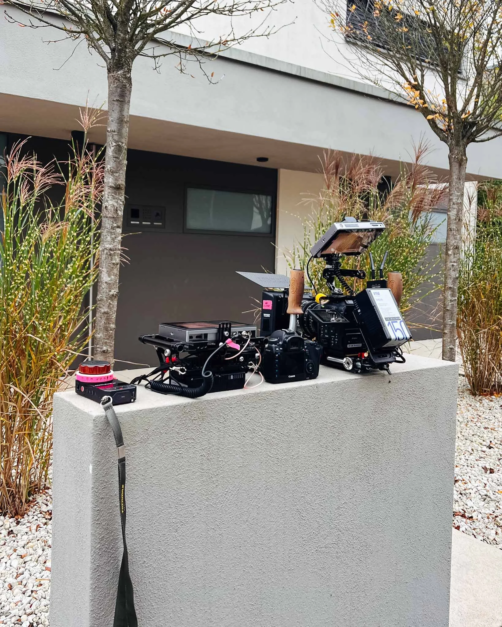 Equipment setup on a white concrete block outdoors, including cameras, monitors, batteries, and wires, with trees and a modern building in the background.