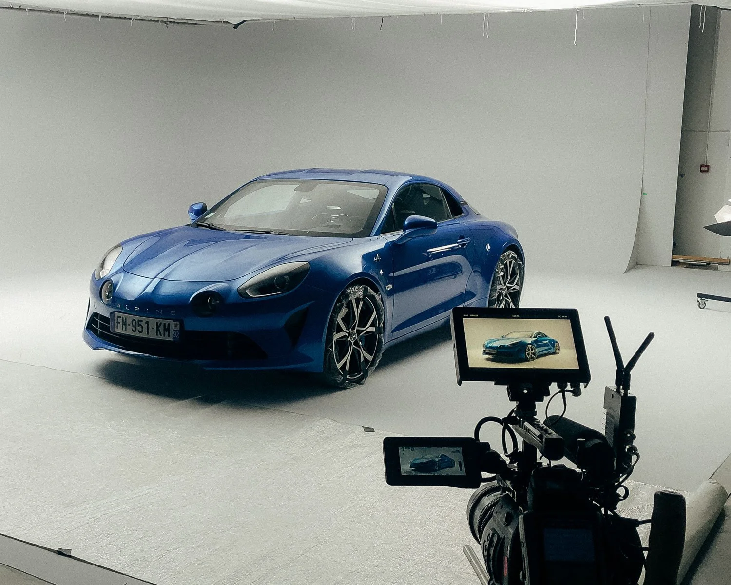 Blue sports car on a white studio backdrop, shot during a professional photoshoot with a camera and monitor visible in the foreground.