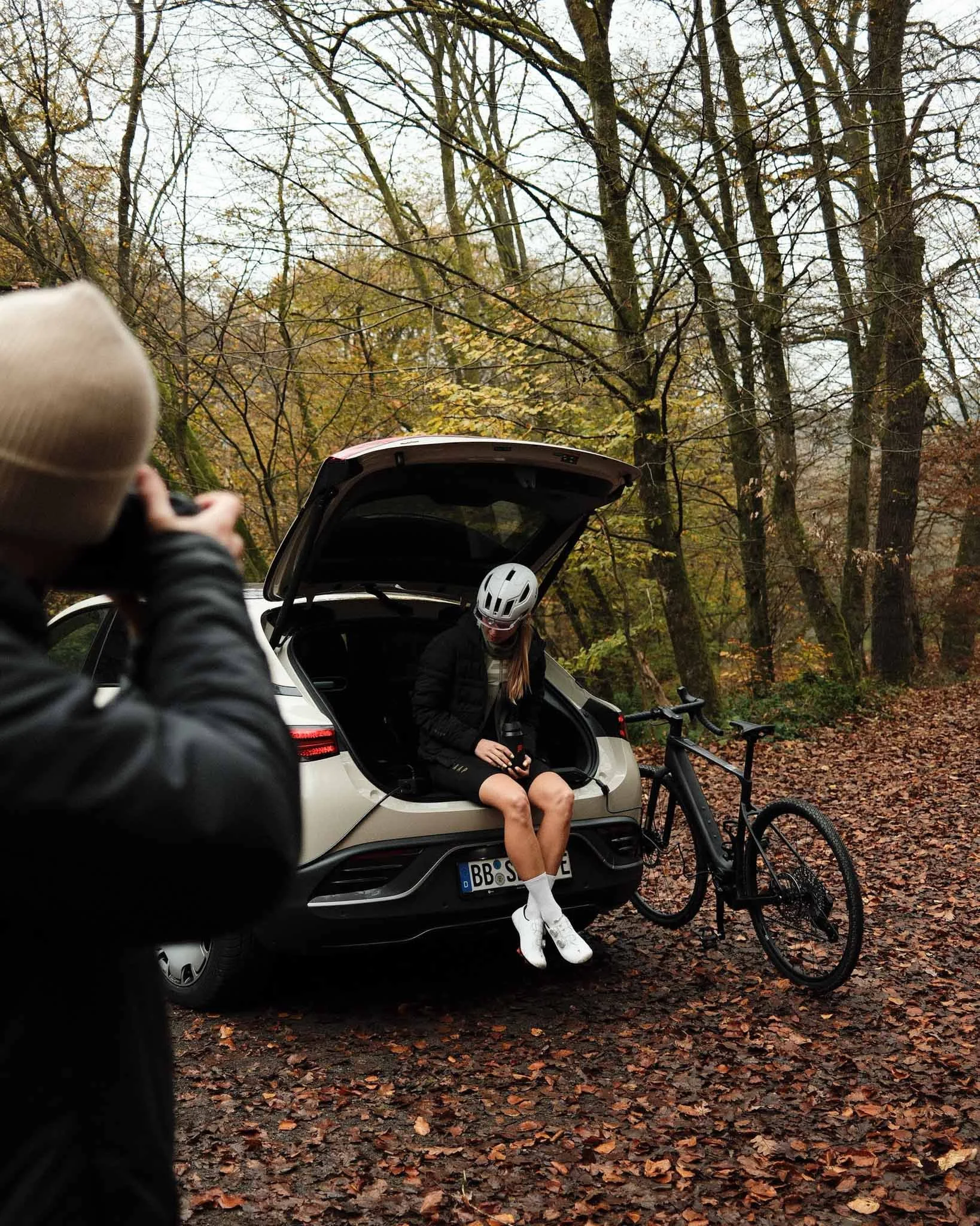 A woman sitting on the open trunk of a white SUV with a black bike leaning against it. She is wearing a white helmet, black jacket, black shorts, and white shoes, and is holding a drink. A person in the foreground is taking her photo. The scene is se