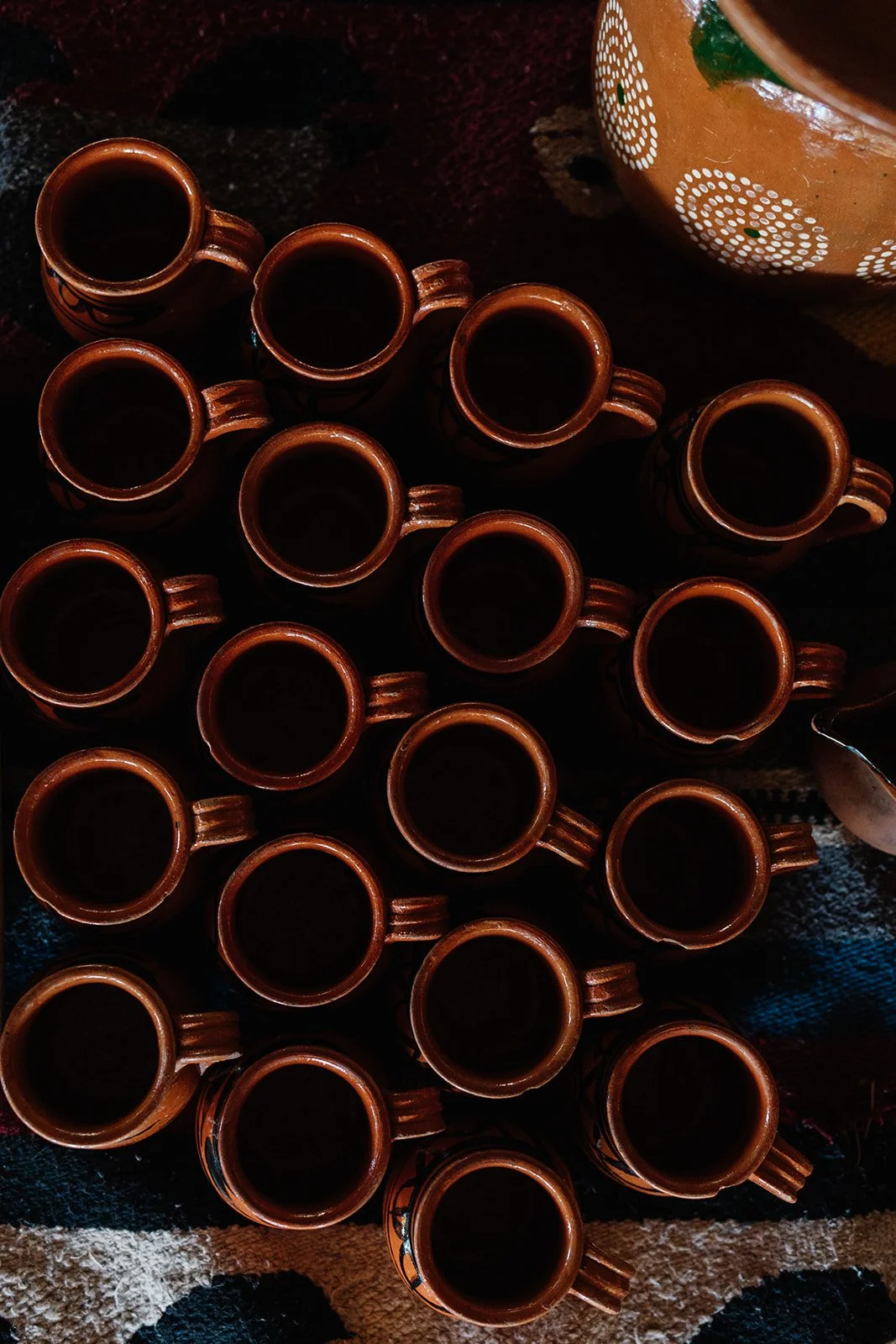 Multiple small brown ceramic cups filled with dark liquid arranged on a dark surface, partly visible larger ceramic bowl with decorative patterns in the background.