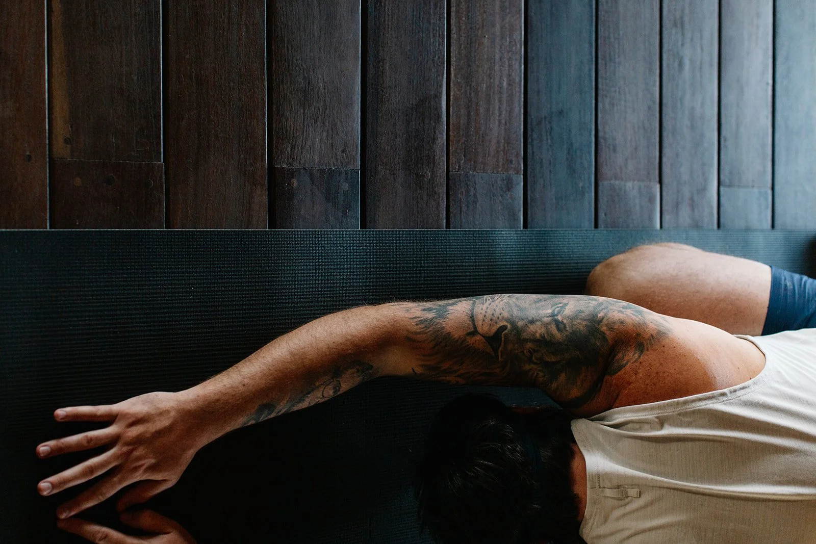 Person practicing yoga or stretching on a black mat against a wooden wall, wearing a white tank top.