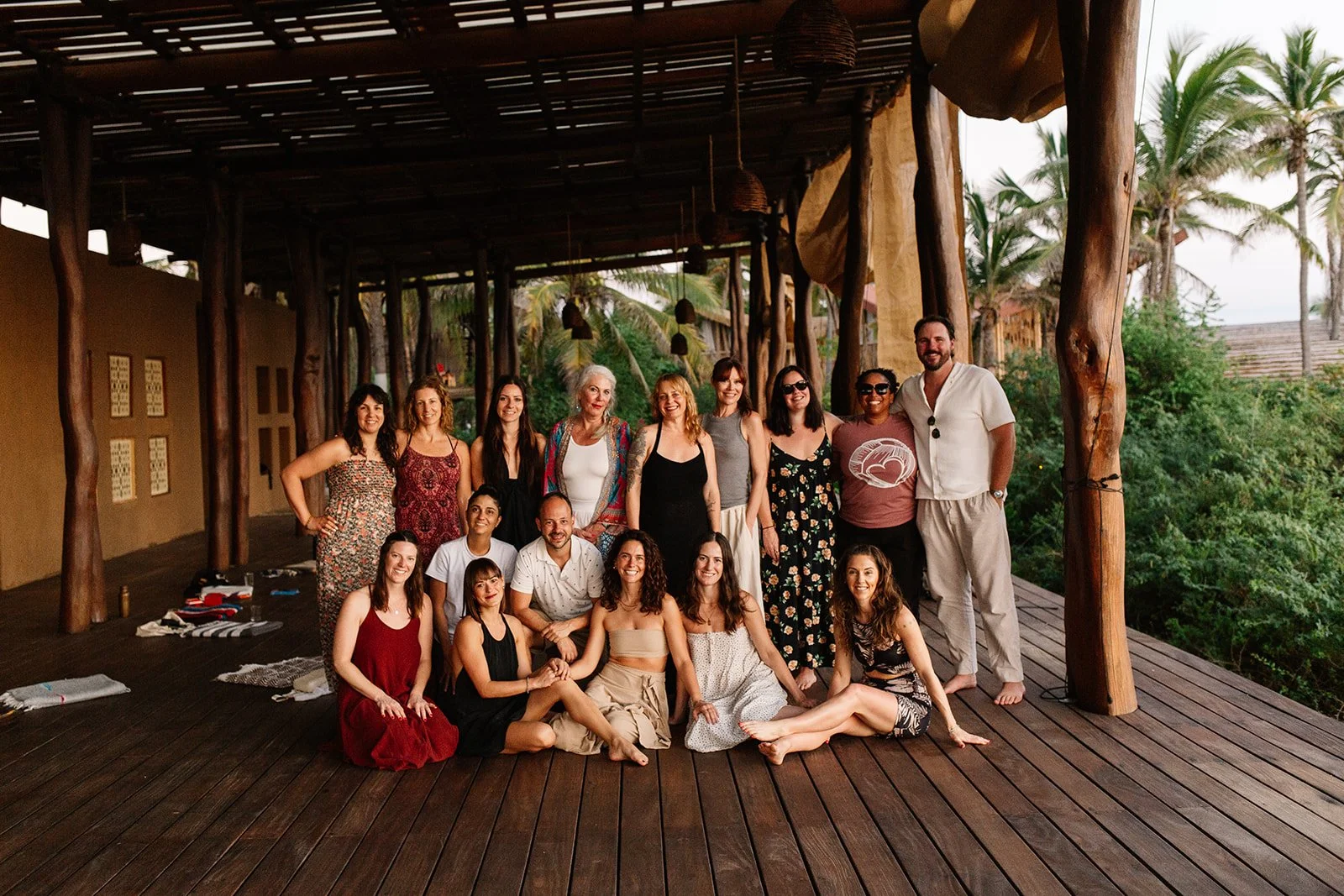 A group of 16 people, mostly women, posing on a wooden deck with tropical trees in the background.