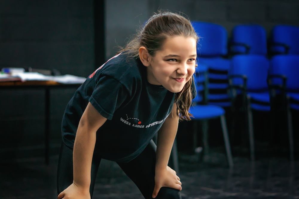 Young girl leaning forward with hands on knees, smiling in a room with black walls and blue chairs in the background.
