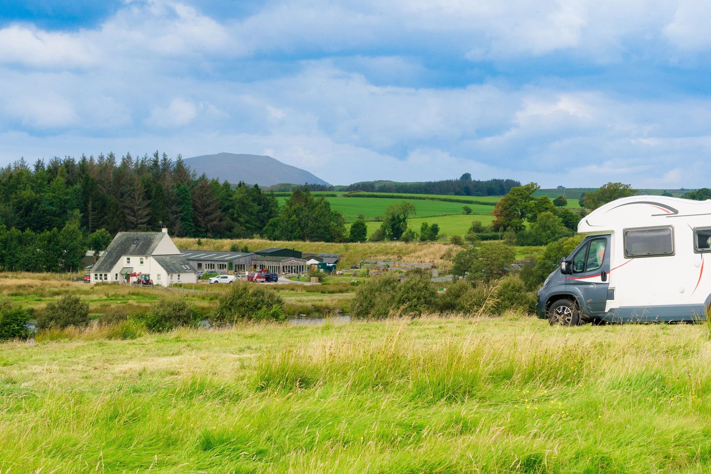 A white RV parked on a grassy field with a rural landscape, trees, and a building in the background under a partly cloudy sky.