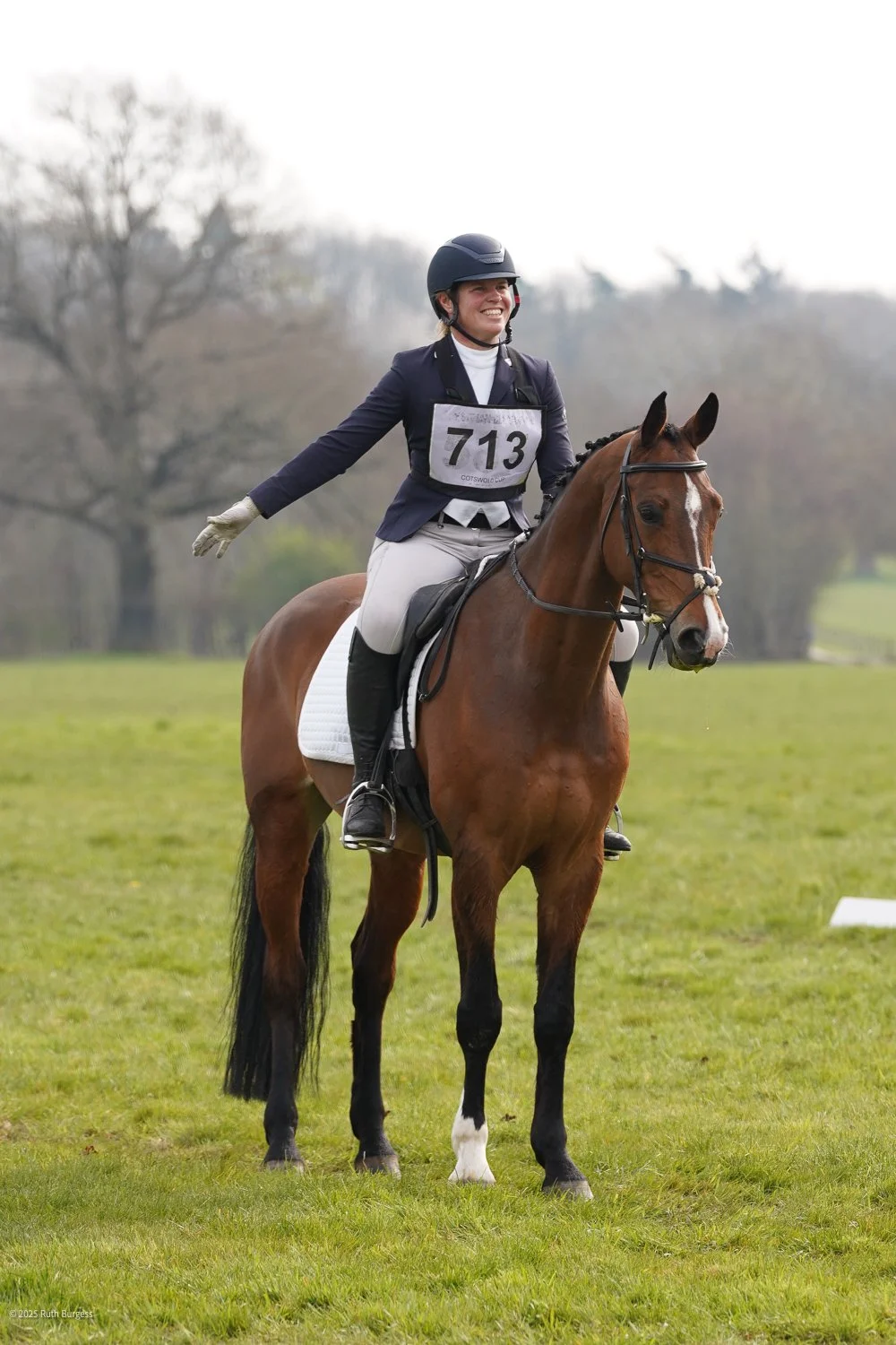 A woman wearing a black riding helmet, beige riding pants, and a navy blazer, sitting on a brown horse with a white marking on its face, during a riding competition outdoors with trees in the background.