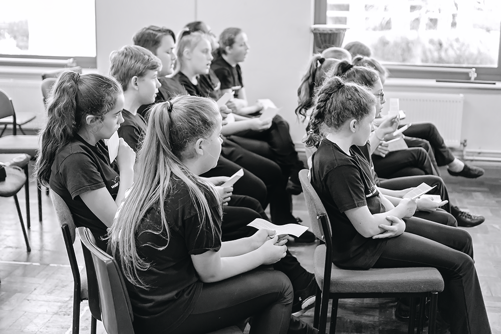 Group of young students sitting in chairs, attentively reading handouts during a class or workshop in a bright room with windows.