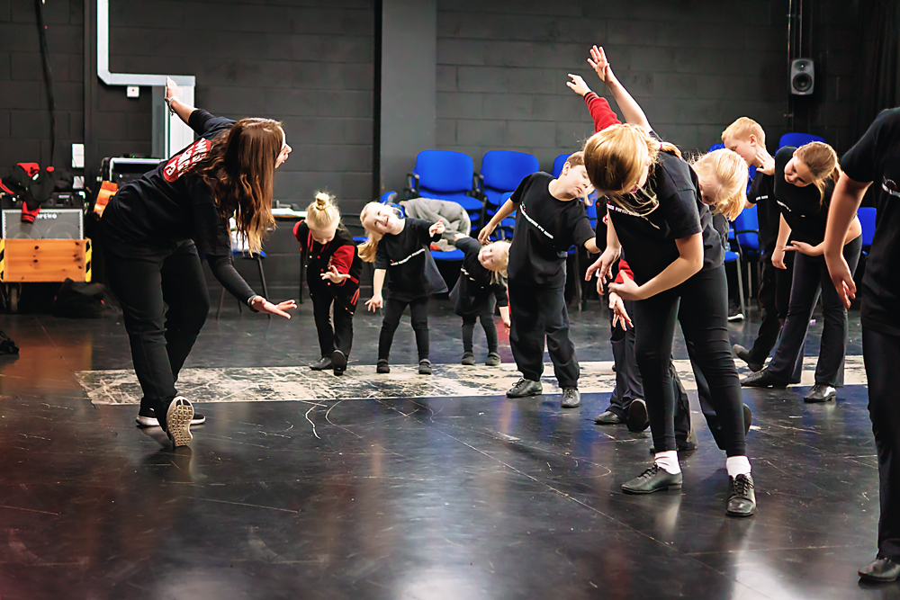 Children and an instructor participating in a dance  class in a rehearsal space with black walls and blue chairs.