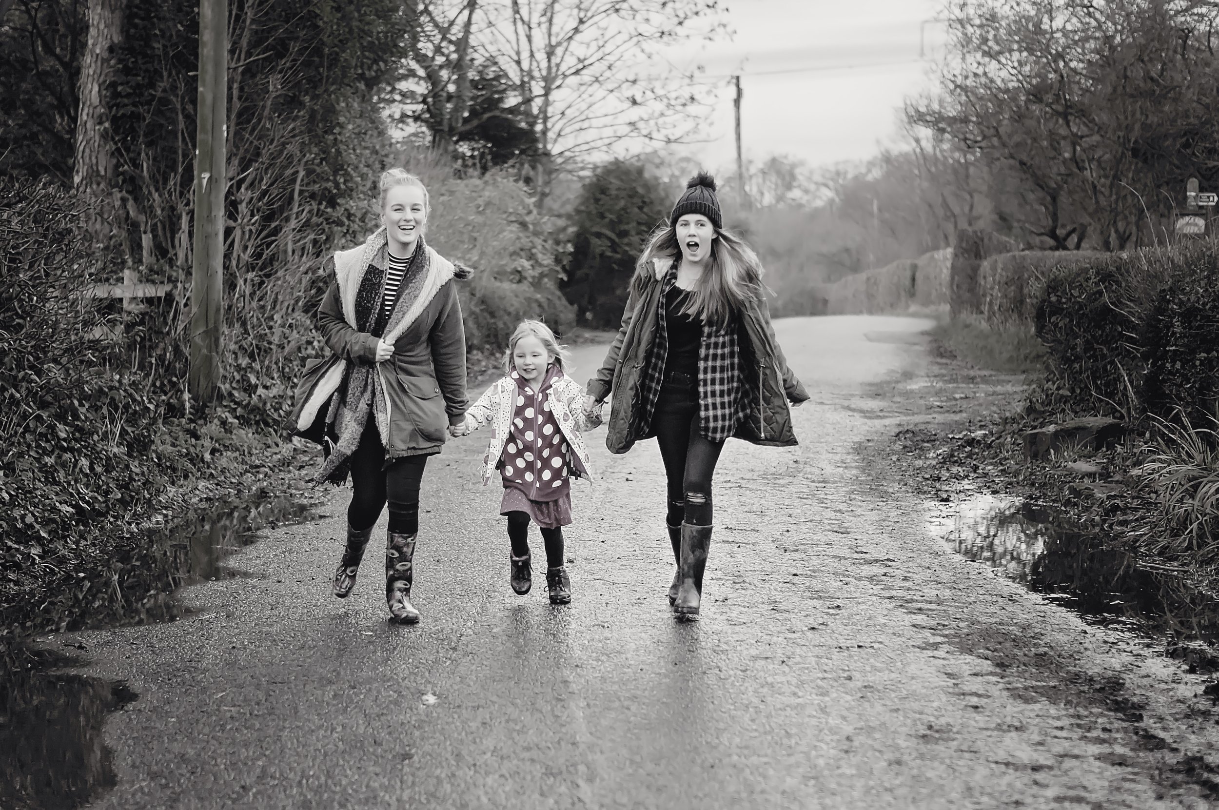 Three females, two young women and one girl, walking on a rural road, holding hands, smiling, and enjoying the outdoors. The women are wearing outdoor jackets and boots; the girl is in a polka-dot jacket.