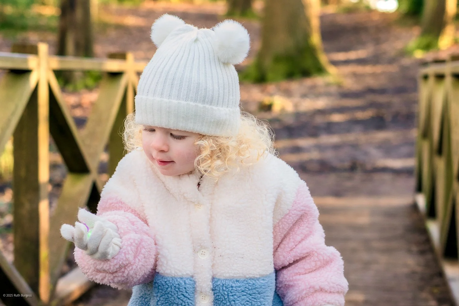 A young girl with curly blonde hair wearing a white hat with bear ears, a pastel-colored fleece jacket, and gloves, standing on a wooden bridge in a park during fall.