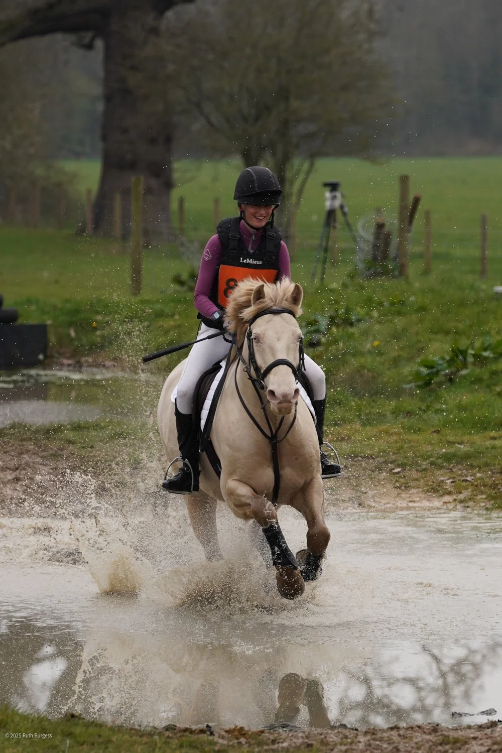 A woman riding a beige horse through a water obstacle during an equestrian event, with trees and fencing in the background.