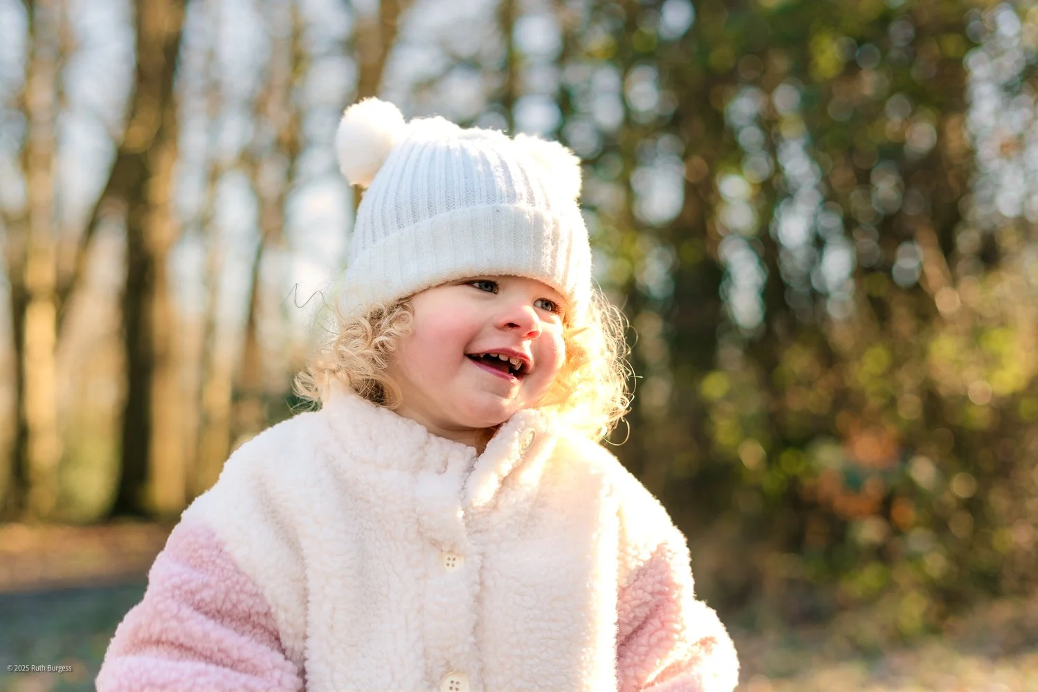 A young girl with curly blonde hair wearing a white knit hat with pom-poms and a fluffy cream-colored coat, smiling outdoors on a sunny day with trees in the background.