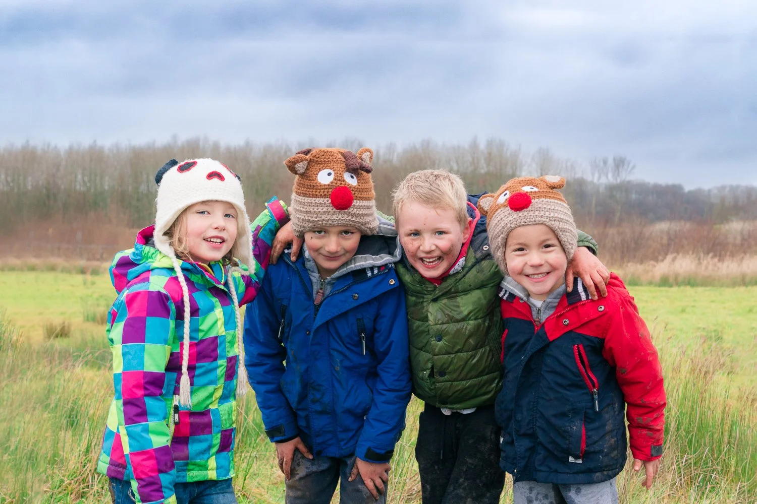 Four children standing together outdoors in a grassy field, wearing colorful jackets and knit hats with reindeer and panda faces, smiling at the camera on a cloudy day.