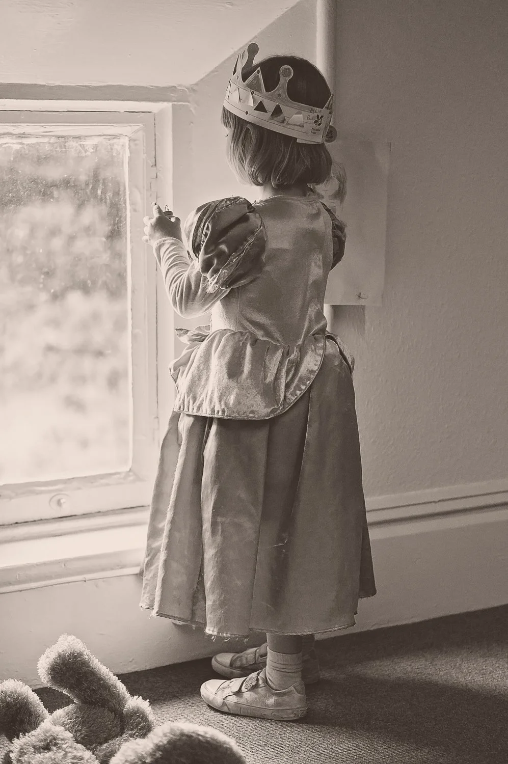 A young girl with a crown on her head, standing by a window inside a house, looking outside. She is wearing a dress with puffy sleeves, sneakers, and socks.