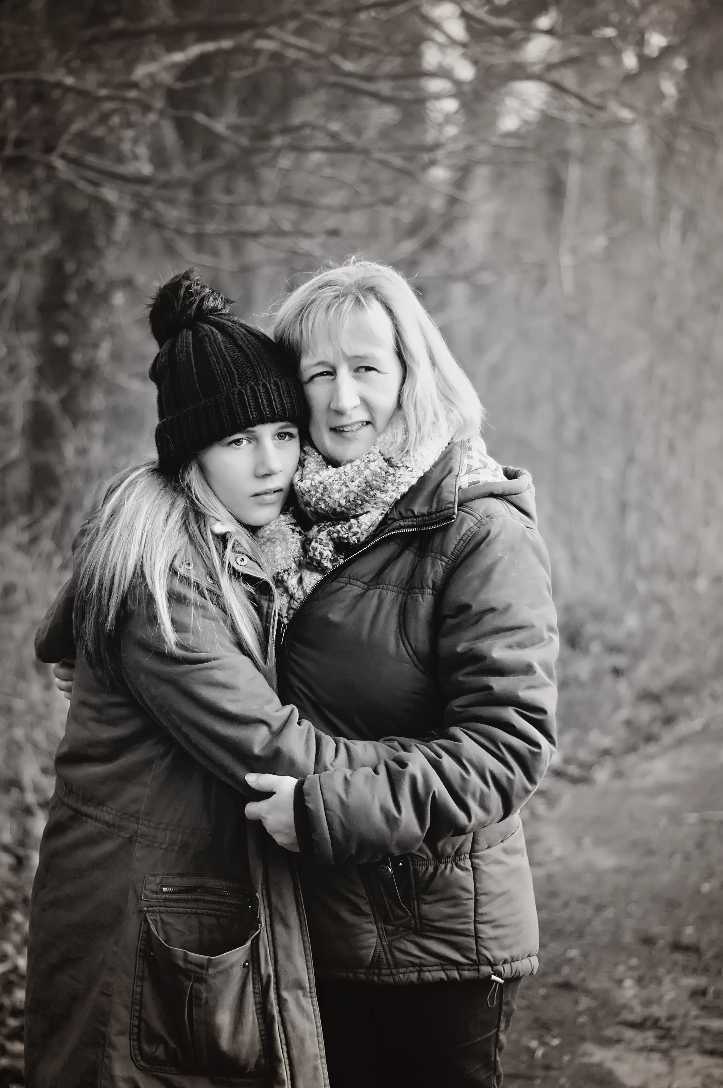 A woman and a girl hugging outdoors, both wearing winter jackets and scarves, with the woman also wearing a knit hat, in a wooded area.