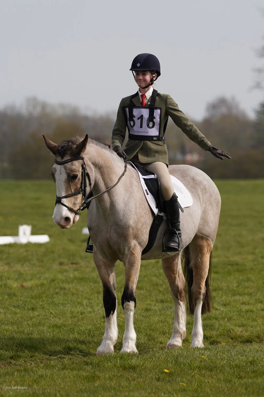 A young female rider in formal riding attire, with a black helmet and gloves, sitting on a tan and white horse in a grassy field. She is participating in a riding competition, wearing a bib with the number 516, and appears to be balancing with her ar