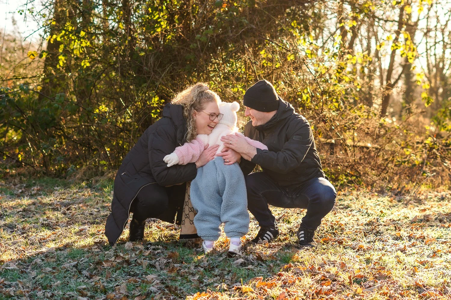 A couple and their young child outdoors in a wooded area during fall, smiling and playing together with the child as the center of attention.