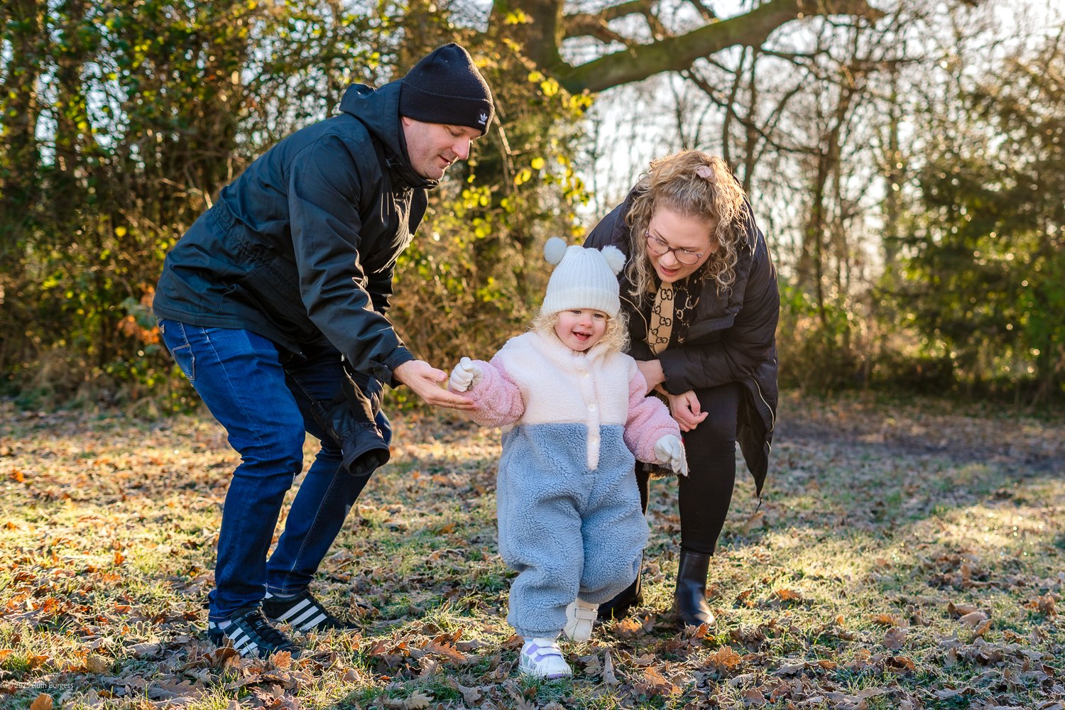 A young girl in a fleece snow suit and white hat being supported by two adults, a man and a woman, as she walks outdoors through fallen leaves in a wooded area during daytime.