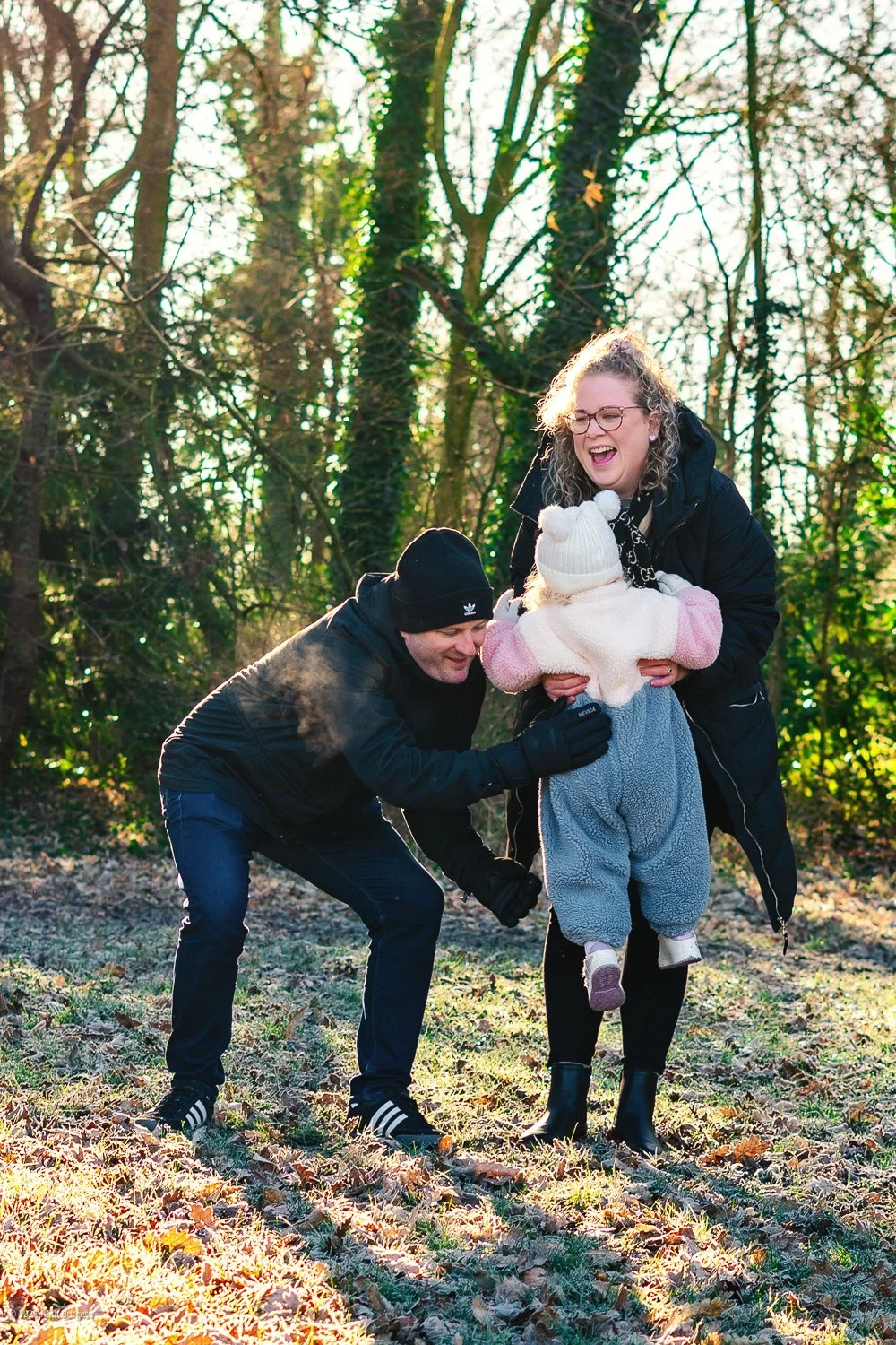 A family of three, including a woman with curly hair and glasses, a man dressed warmly, and a young girl in a hat, are playing and laughing together in a wooded outdoor area during sunlight.