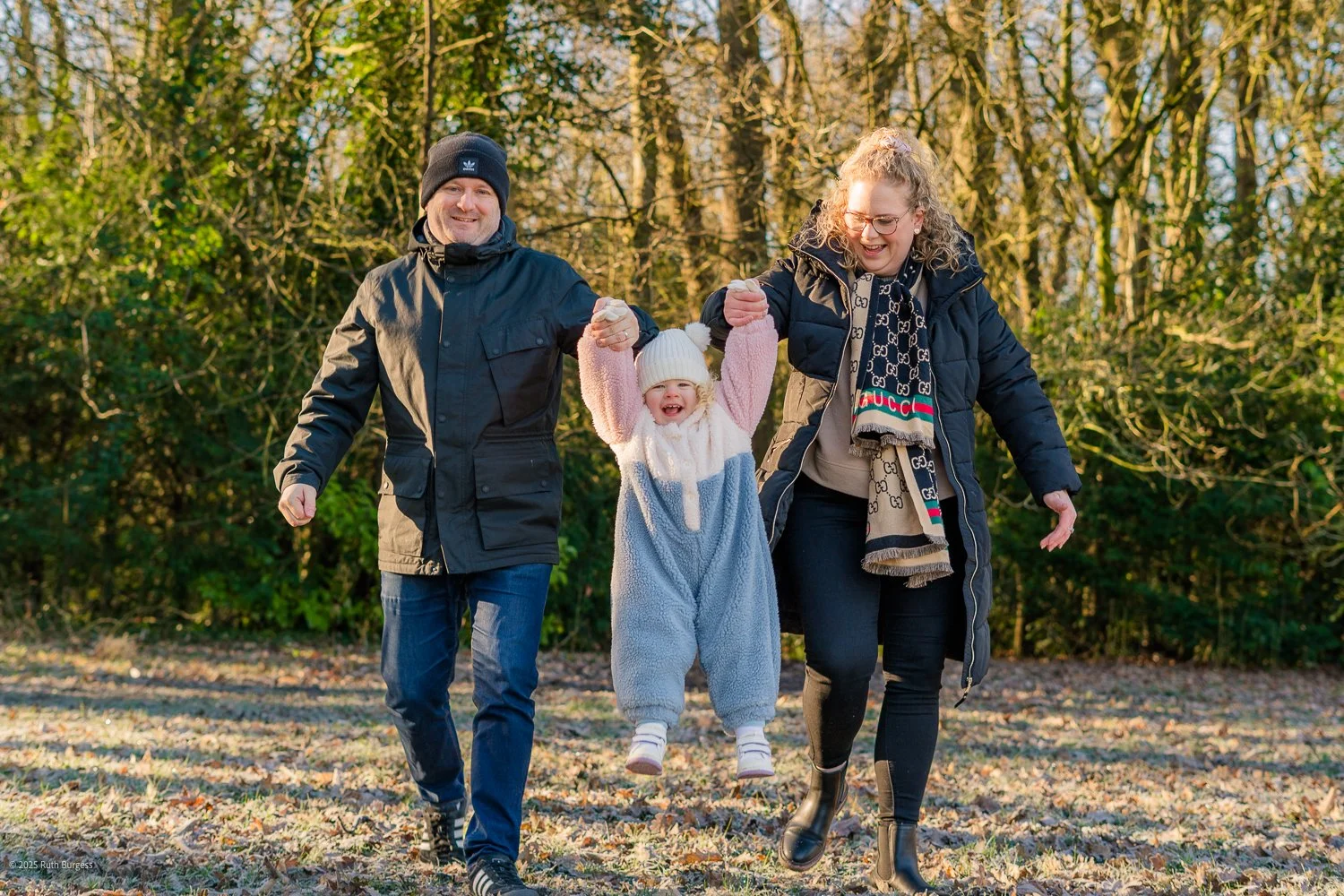 A happy family of three, including a young girl in a cozy onesie, being lifted in the air by her parents while walking outdoors in a wooded area during daytime.
