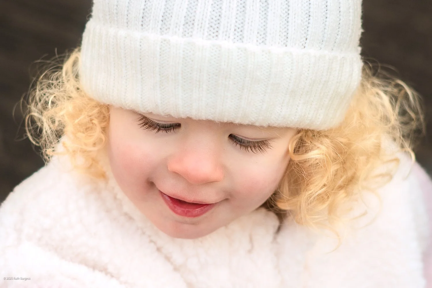 Close-up of a young girl with curly blonde hair wearing a white knit hat and a white sweater, smiling softly with her eyes closed.