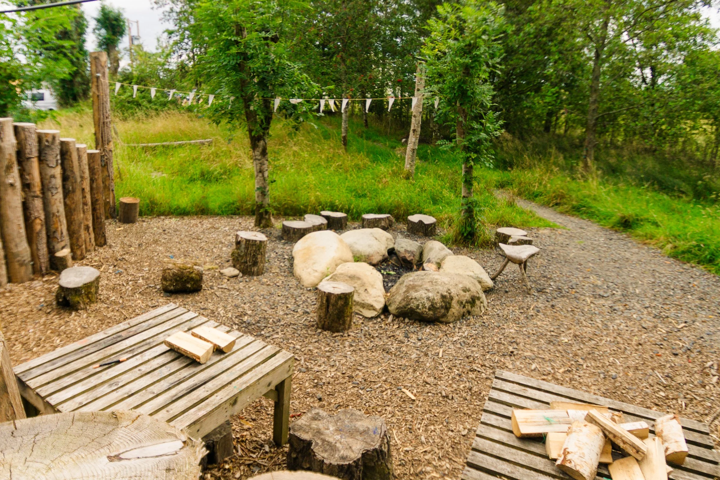 Outdoor fire pit area with rocks, wooden stumps, and benches, surrounded by trees and greenery.