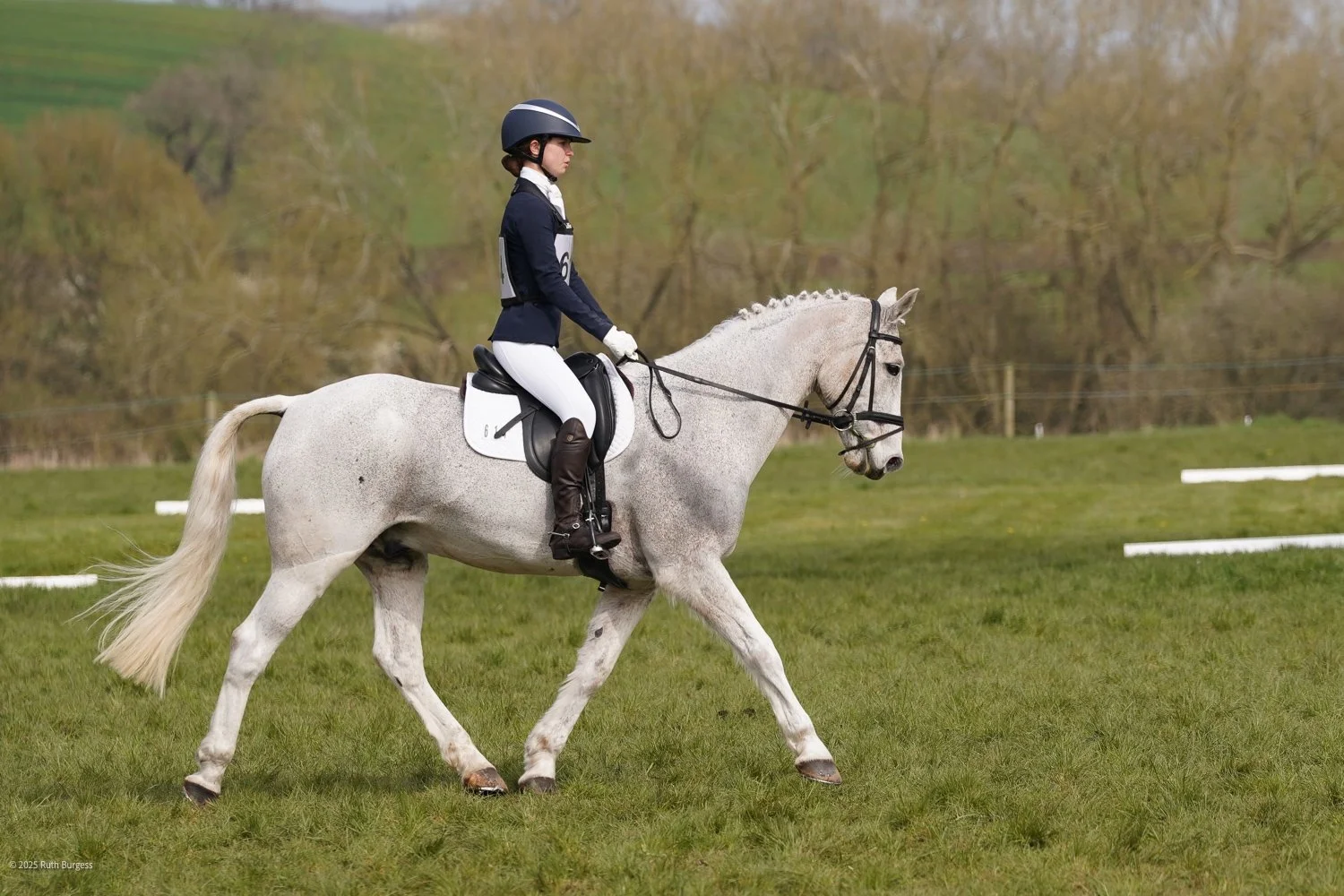 A woman in equestrian riding attire riding a white horse during a competition on a grassy field.
