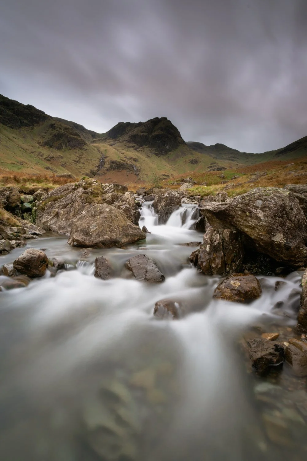A mountain stream with rocks flowing through a green valley under cloudy skies.