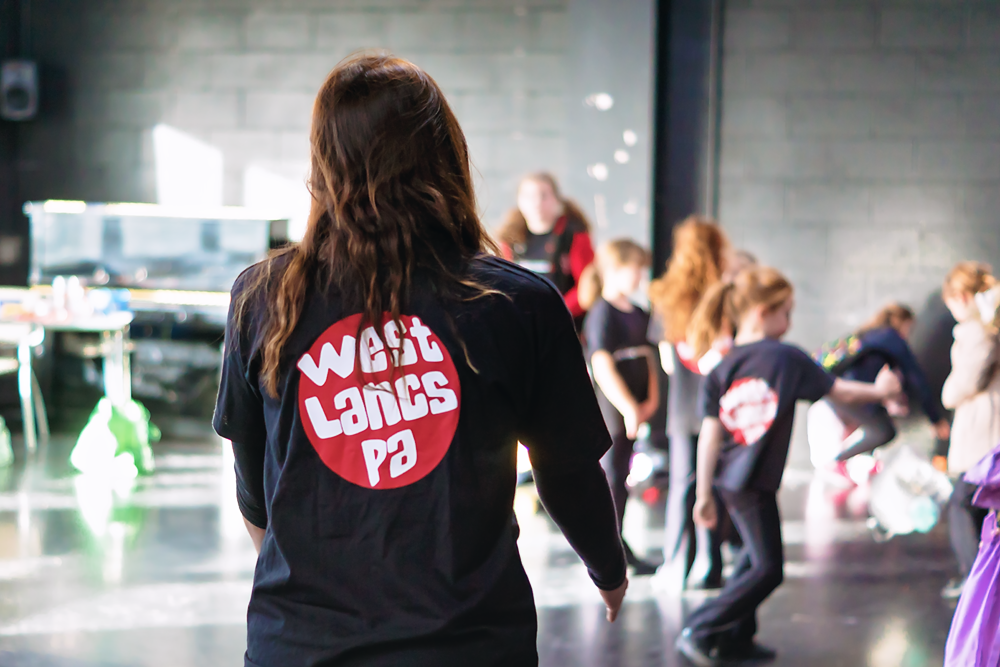 Back of a woman with long brown hair wearing a black shirt with a red circular logo that reads 'West Lance Pa' on her back, with children in the background participating in a dance class or activity in a studio with a concrete wall and large windows.