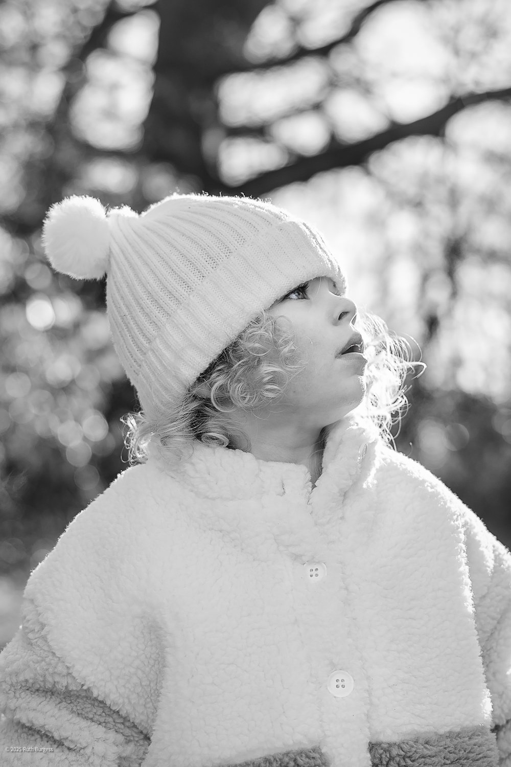 A young child with curly hair, wearing a knit hat with a pom-pom and a fuzzy coat, looks up outdoors in a snowy environment, with blurred trees in the background.